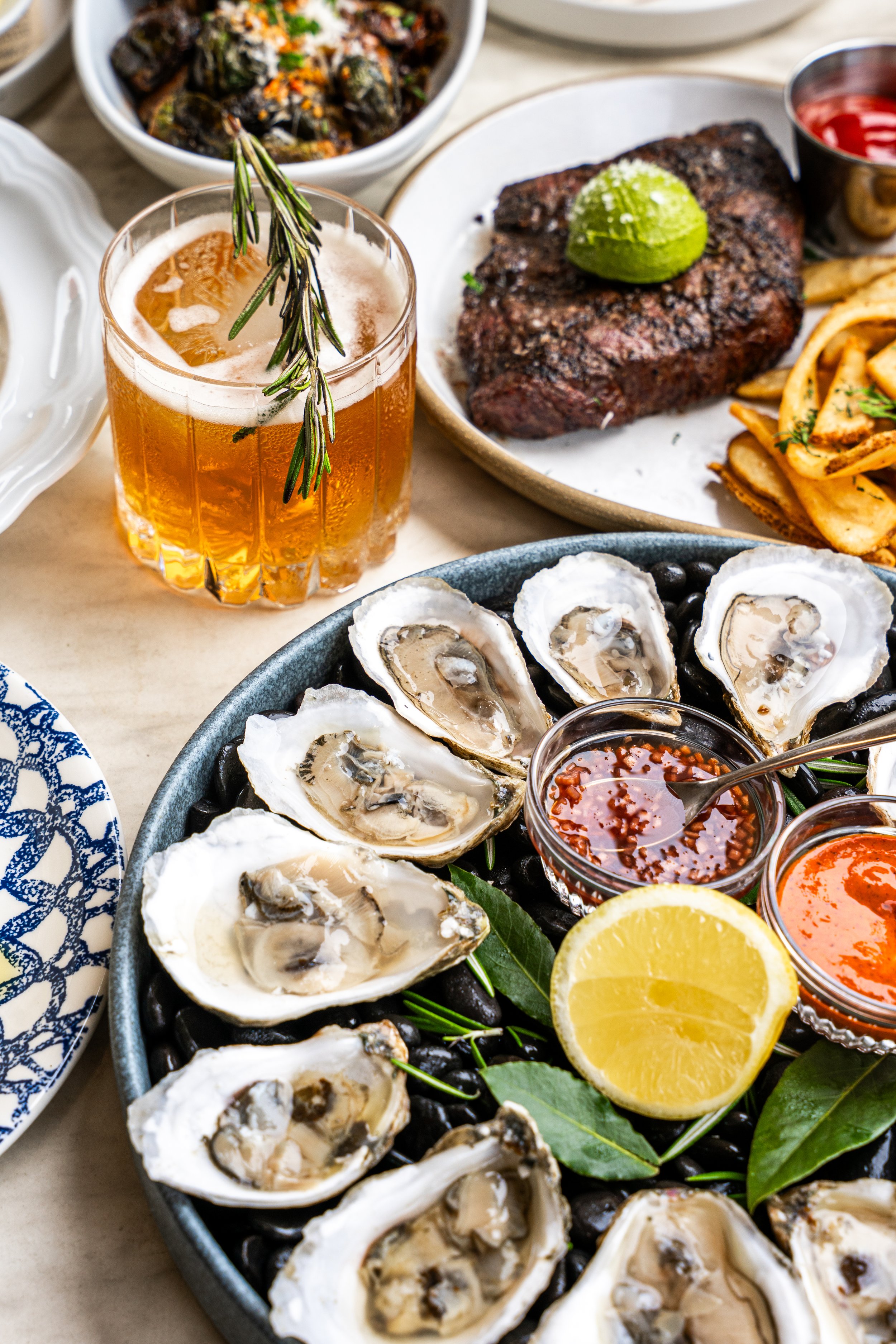 Oysters on the half shell served with lemon wedges, cocktail sauces, and garnishes at a seafood restaurant. There is a drink with rosemary and ice in a glass.