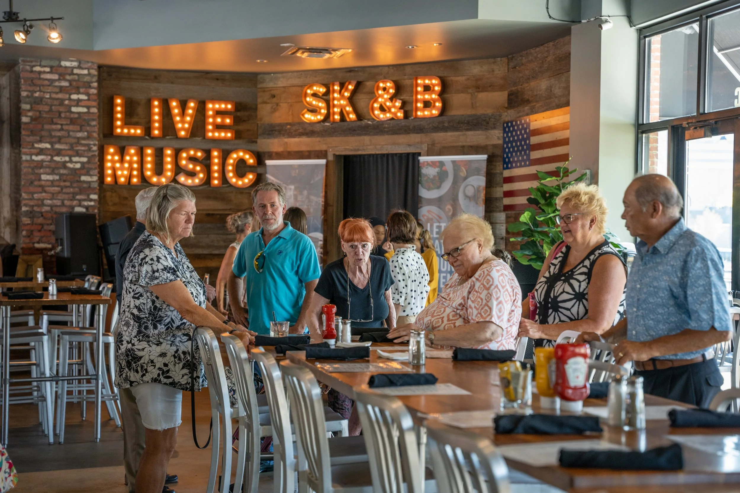 A group of older adults gathered around a restaurant table inside, with some standing and some seated, engaged in conversation, with bright natural light coming from large windows. In the background, there are decorative signs that read 'LIVE MUSIC' 