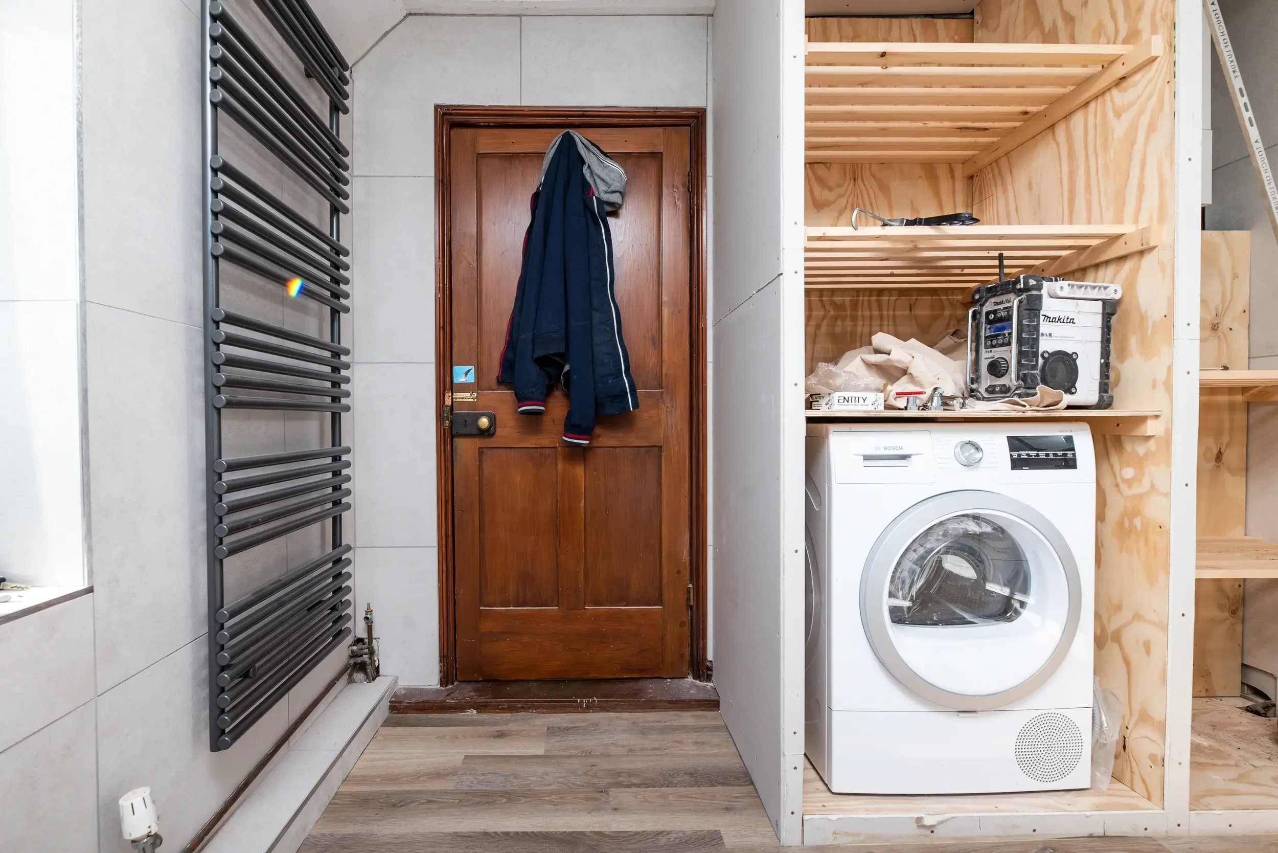 A laundry room with a wooden door, a gray towel hanging on the door, a black radiator on the wall, a washing machine, and a wooden shelf with tools and supplies.