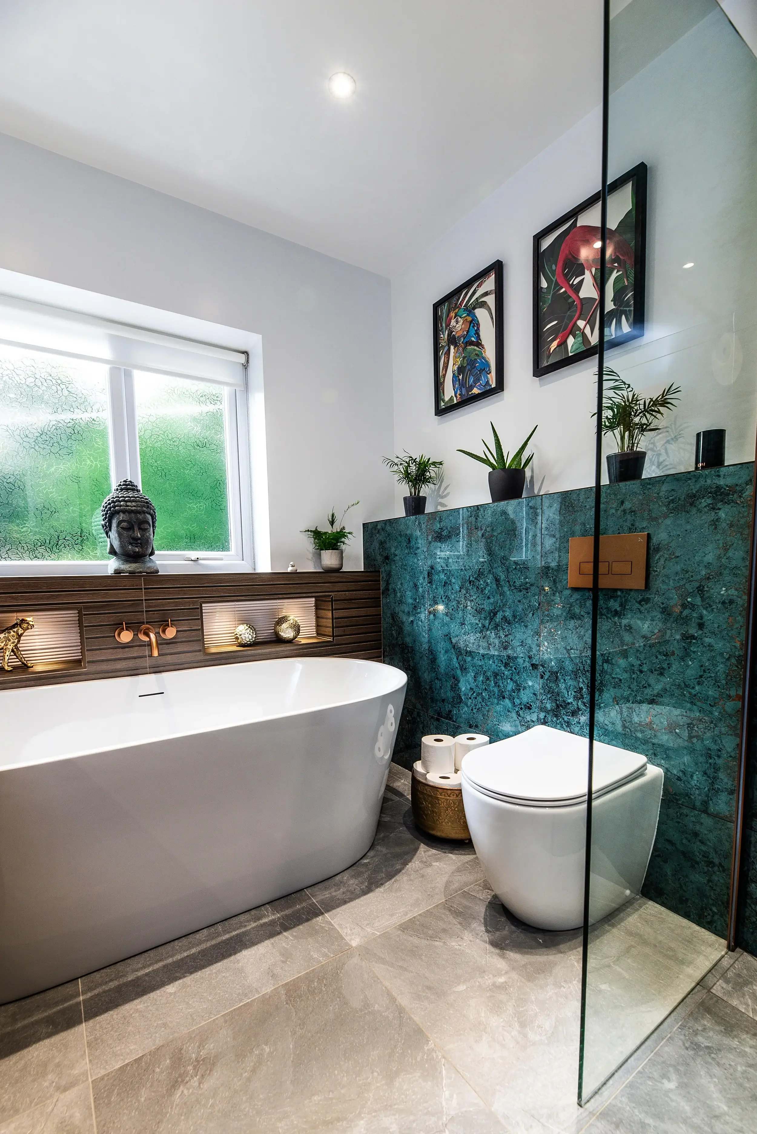 A modern bathroom featuring a white freestanding bathtub, a toilet, a green marble accent wall, indoor potted plants, framed tropical-themed art, a window with frosted glass, and decorative items including a Buddha head sculpture and golden knick-knacks.