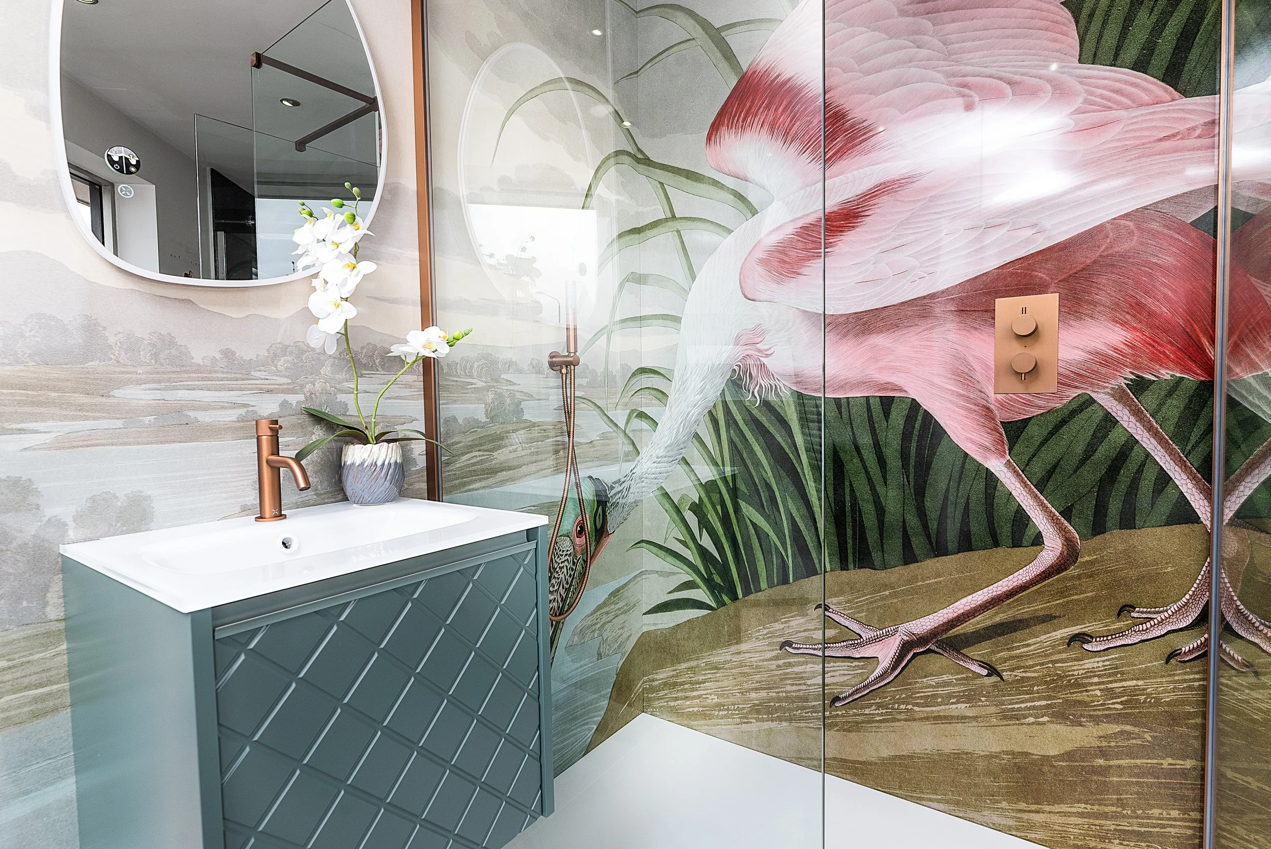 A bathroom with a large flamingo mural on the wall, a small sink with a copper faucet, a decorative vase with white orchids, a wall mirror, and a glass shower door.