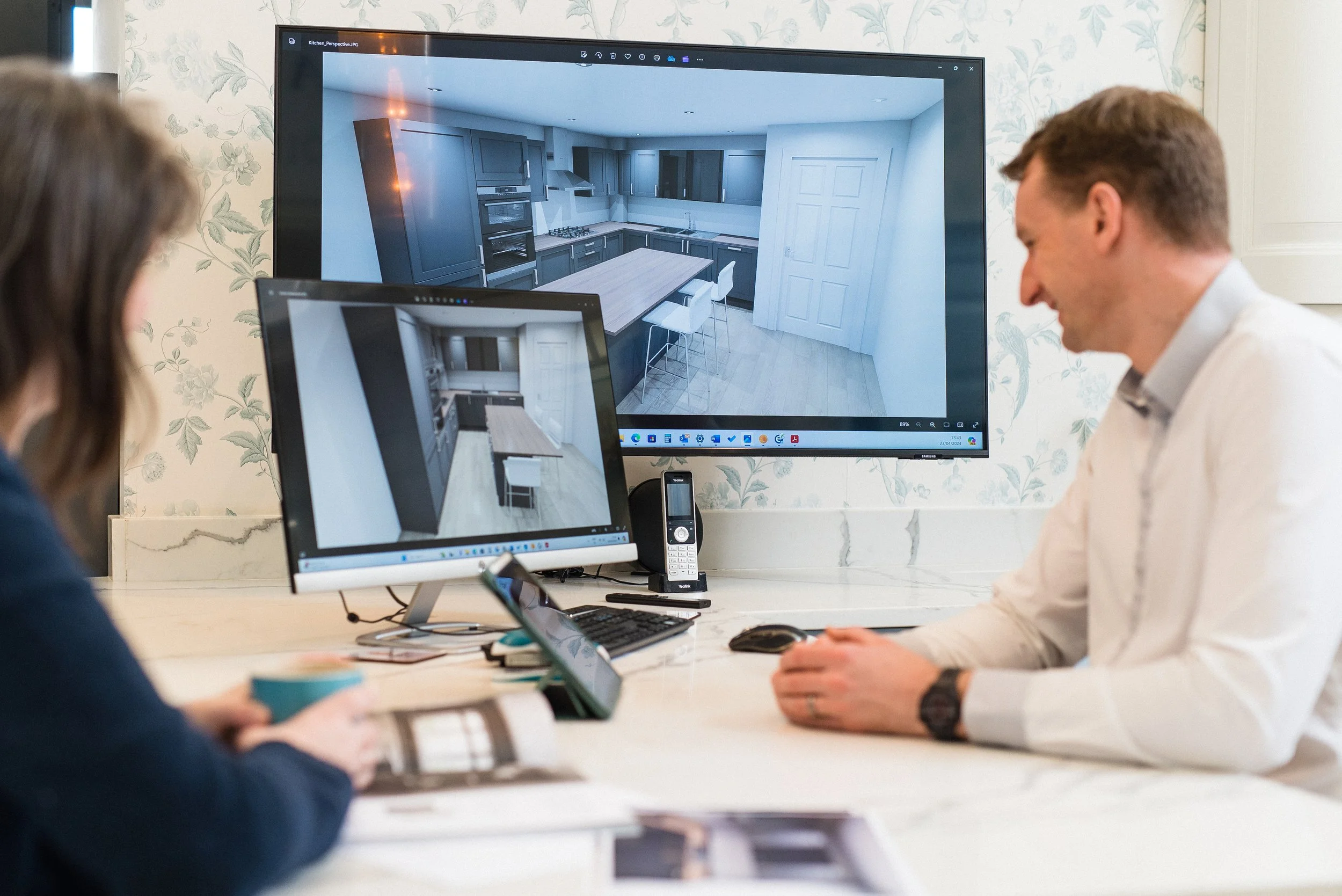 Two people sitting at a desk looking at computer screens displaying a kitchen design.