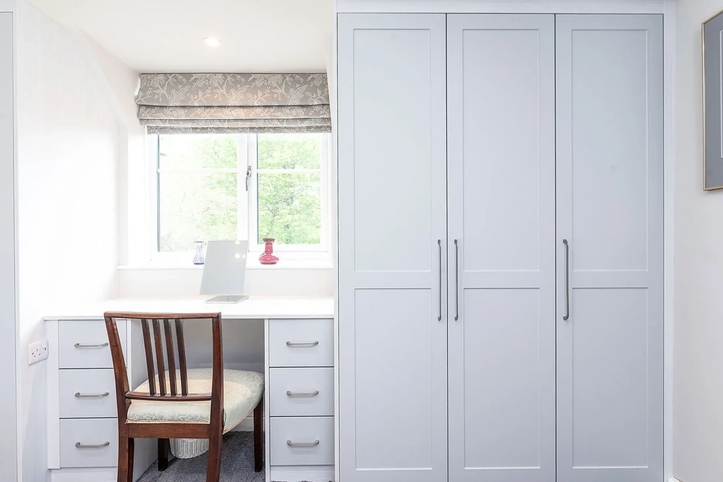 A corner of a room with a built-in desk, a wooden chair with a cushioned seat, a window with floral patterned curtains, a red vase, and tall white cabinets with handles.