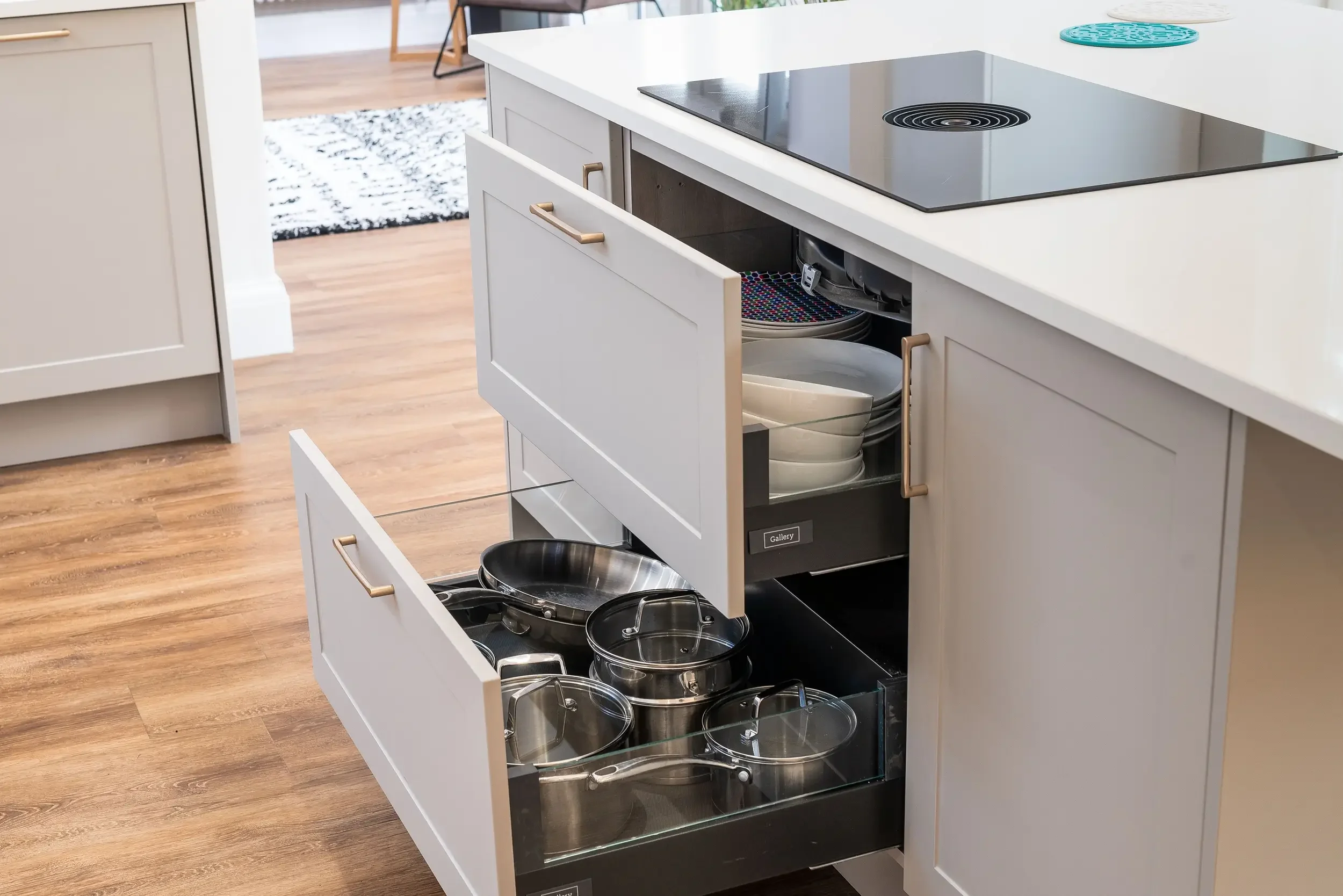 Open kitchen cabinets revealing a pull-out drawer filled with various pots and bowls, with a white countertop and kitchen in the background.