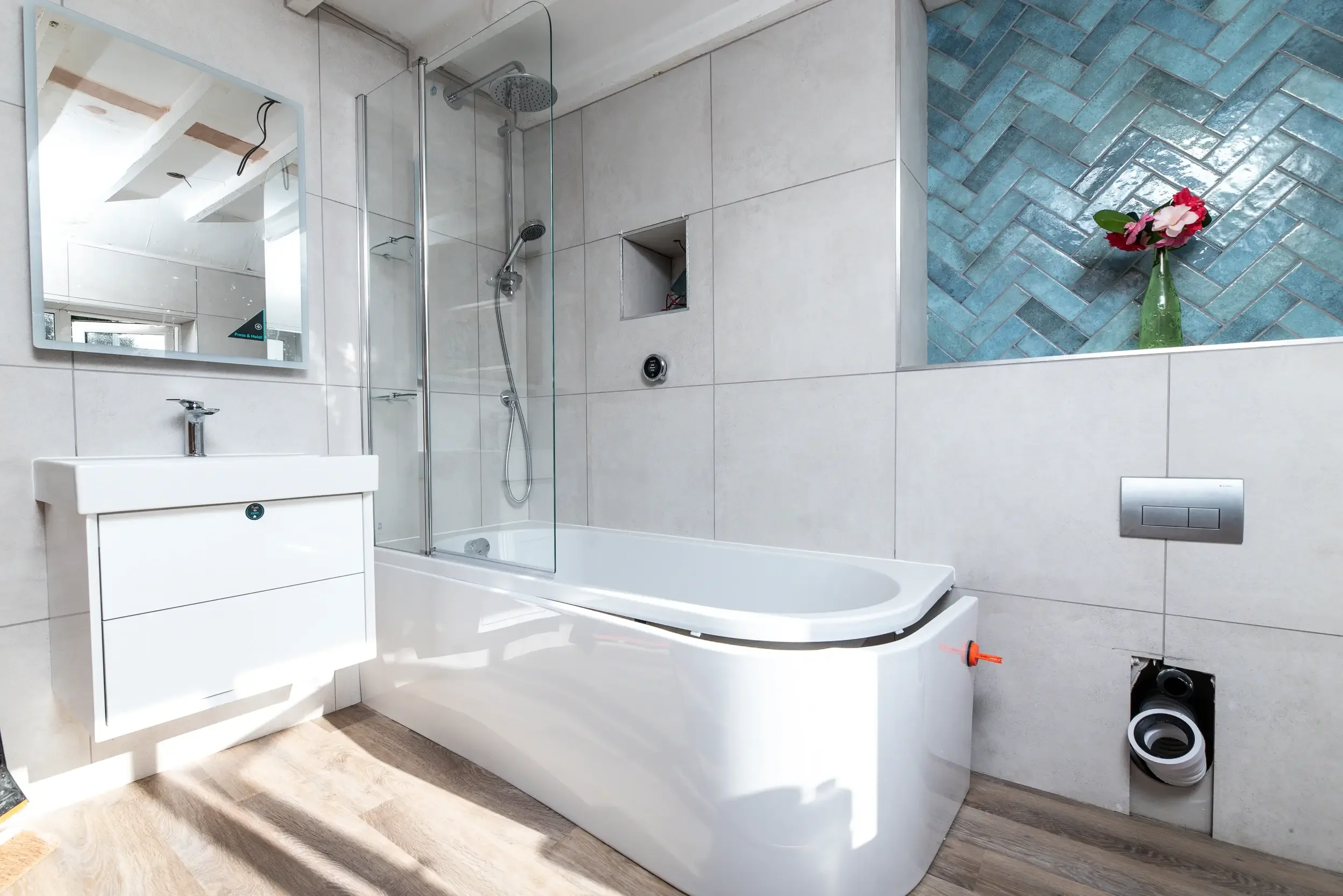 A bathroom under renovation with a white bathtub, a small white sink cabinet, a mirror, and a glass shower enclosure. There's a decorative blue tile wall section with a vase of pink and white flowers. Tools and pipes are visible, indicating ongoing w