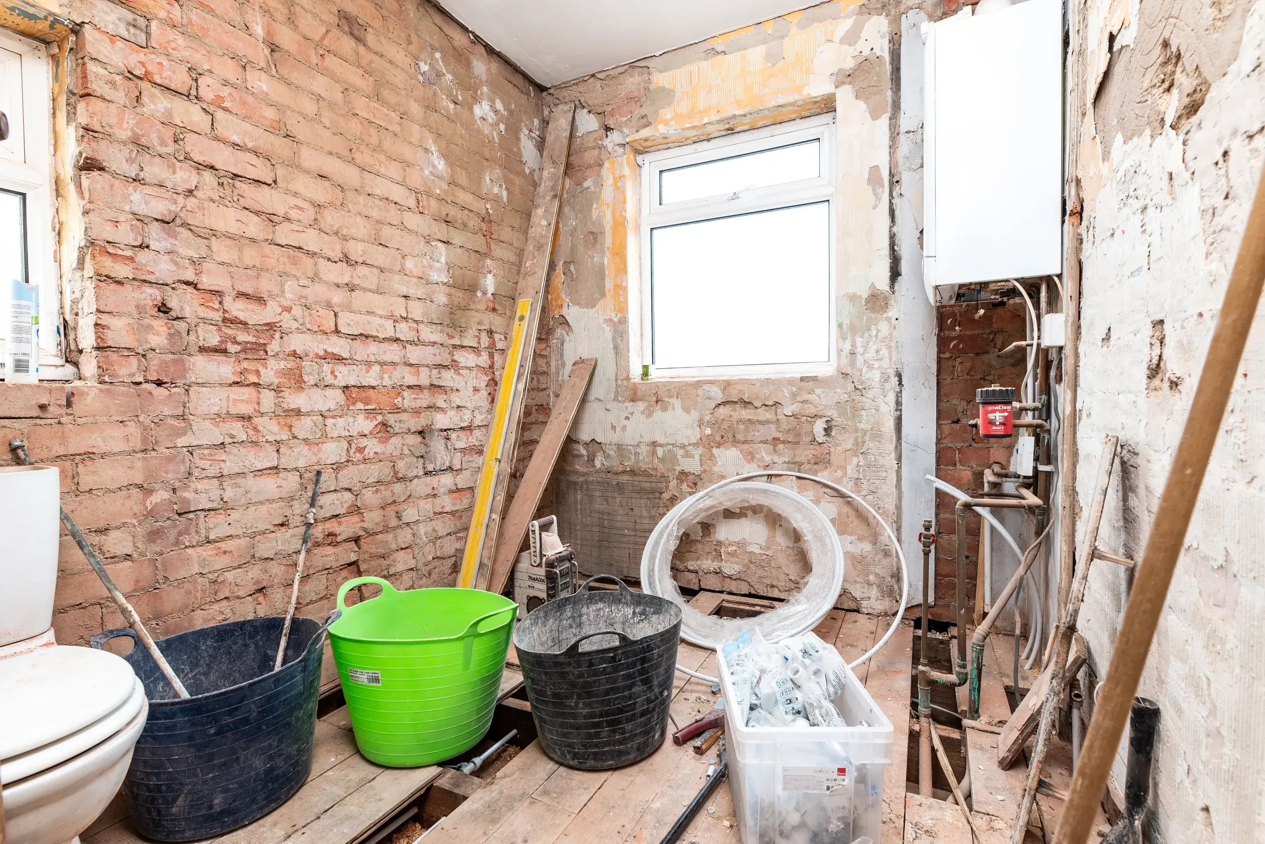 Room under renovation with exposed brick walls, construction tools, and buckets of construction supplies. A window is visible in the background, and various construction materials and equipment are scattered throughout the space.