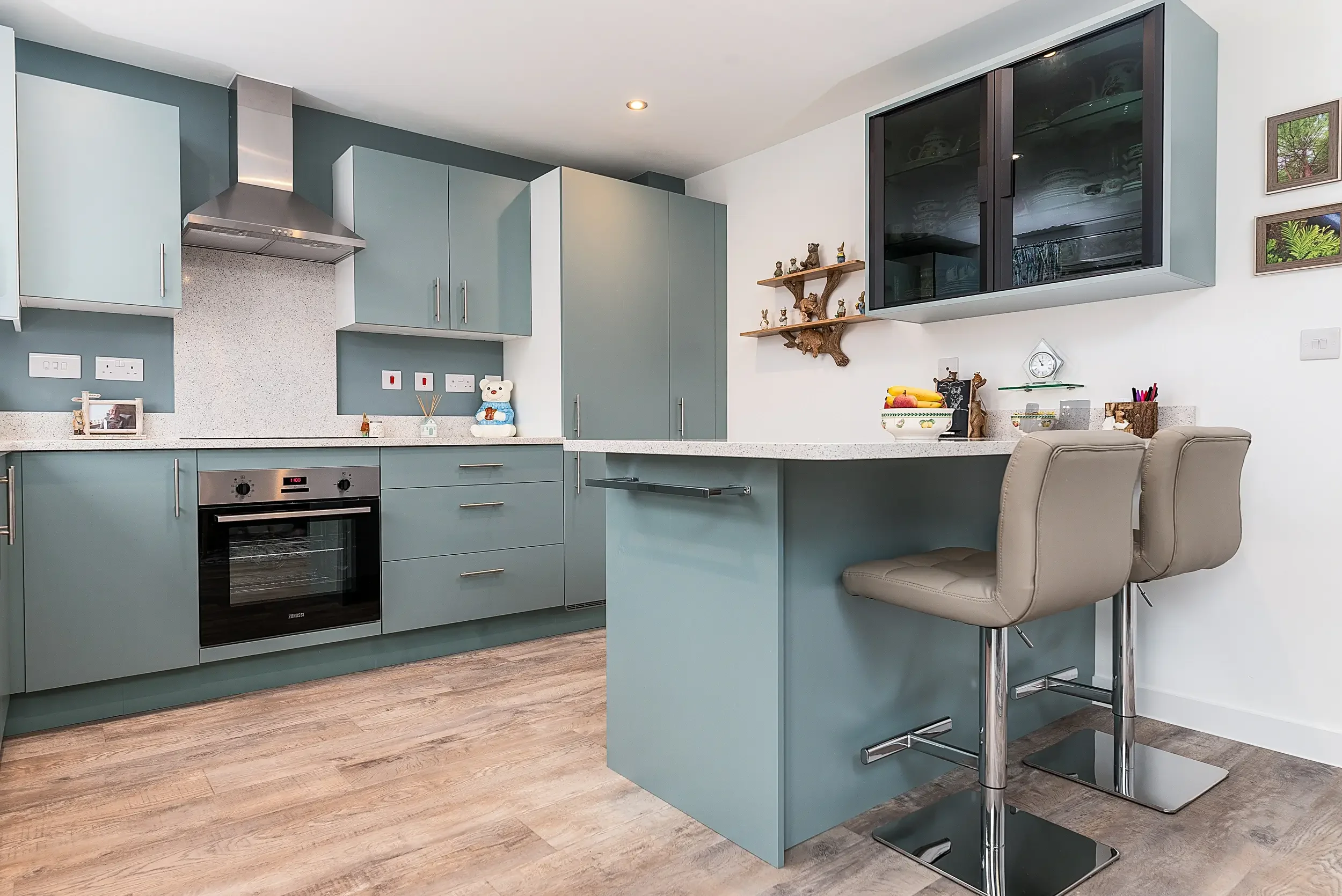 Modern kitchen with pastel blue cabinets, a built-in oven, a grey dining chair, a white wall with small picture frames, and decorative items including a bear figurine and green bowl with bananas.