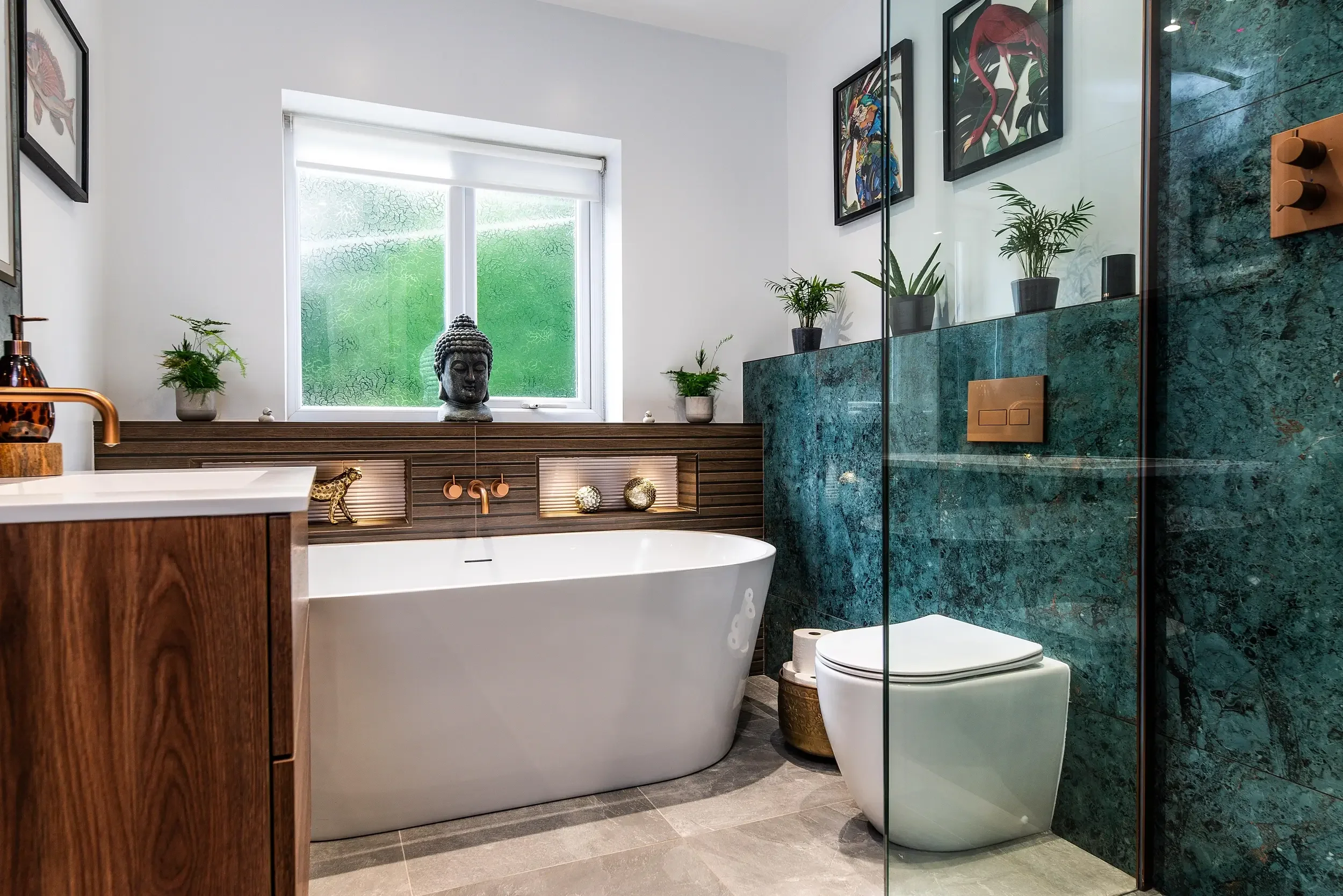 Modern bathroom with a freestanding bathtub, potted plants, framed artwork, green marble wall, toilet, and window with frosted glass.