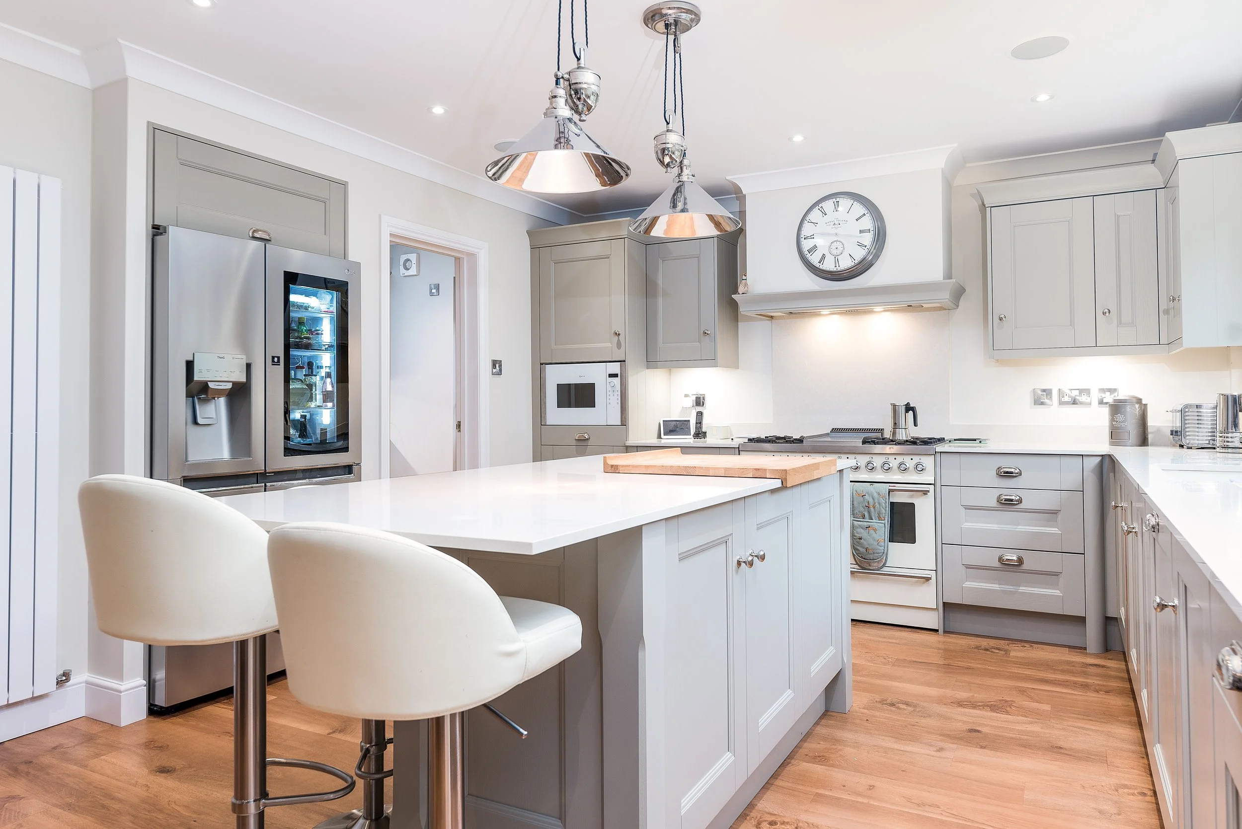 Modern kitchen with white cabinets, a large white island with two beige chairs, stainless steel appliances including a refrigerator and microwave, and a wall clock. Pendant lights hang from the ceiling, and hardwood floors complement the neutral color scheme.