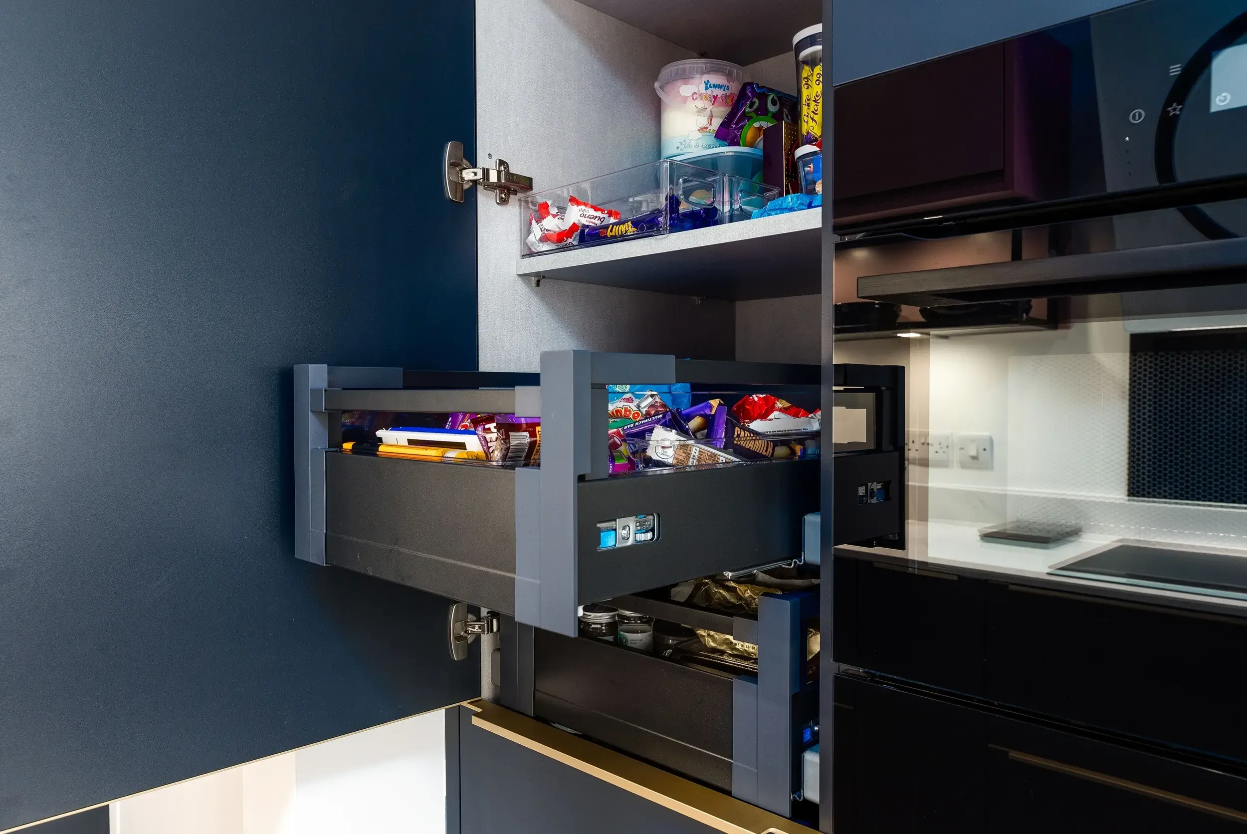 Open kitchen cabinet with snacks and condiments inside, black microwave oven below, dark countertop, and blue wall behind
