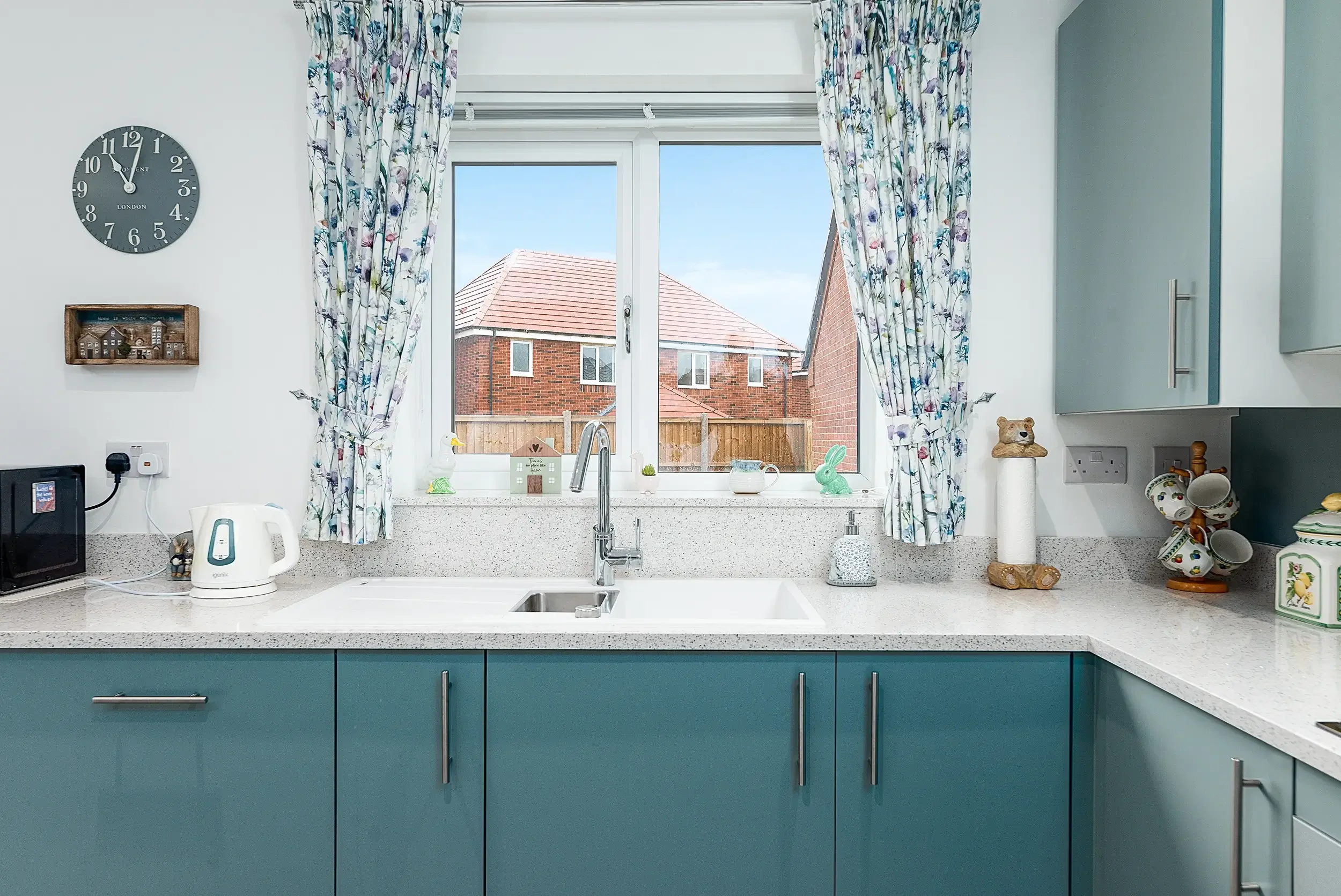 A kitchen sink window with floral curtains, a view of neighboring brick houses, and decorative items on the countertop including a bunny figurine, a small house, and a tea set.