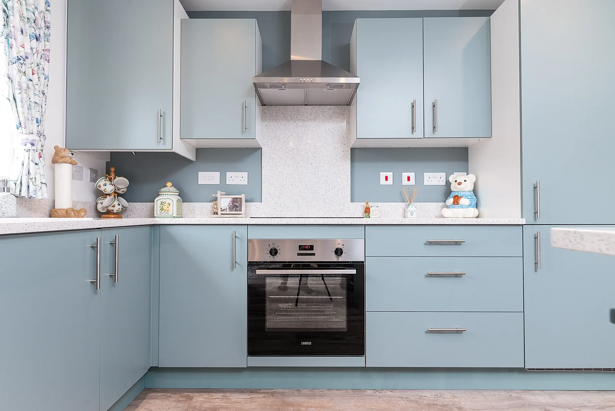 Kitchen with light blue cabinets, a stainless steel oven with control knobs, a range hood above the stove, decorative appliances and items on the countertop, and a window with floral curtains.