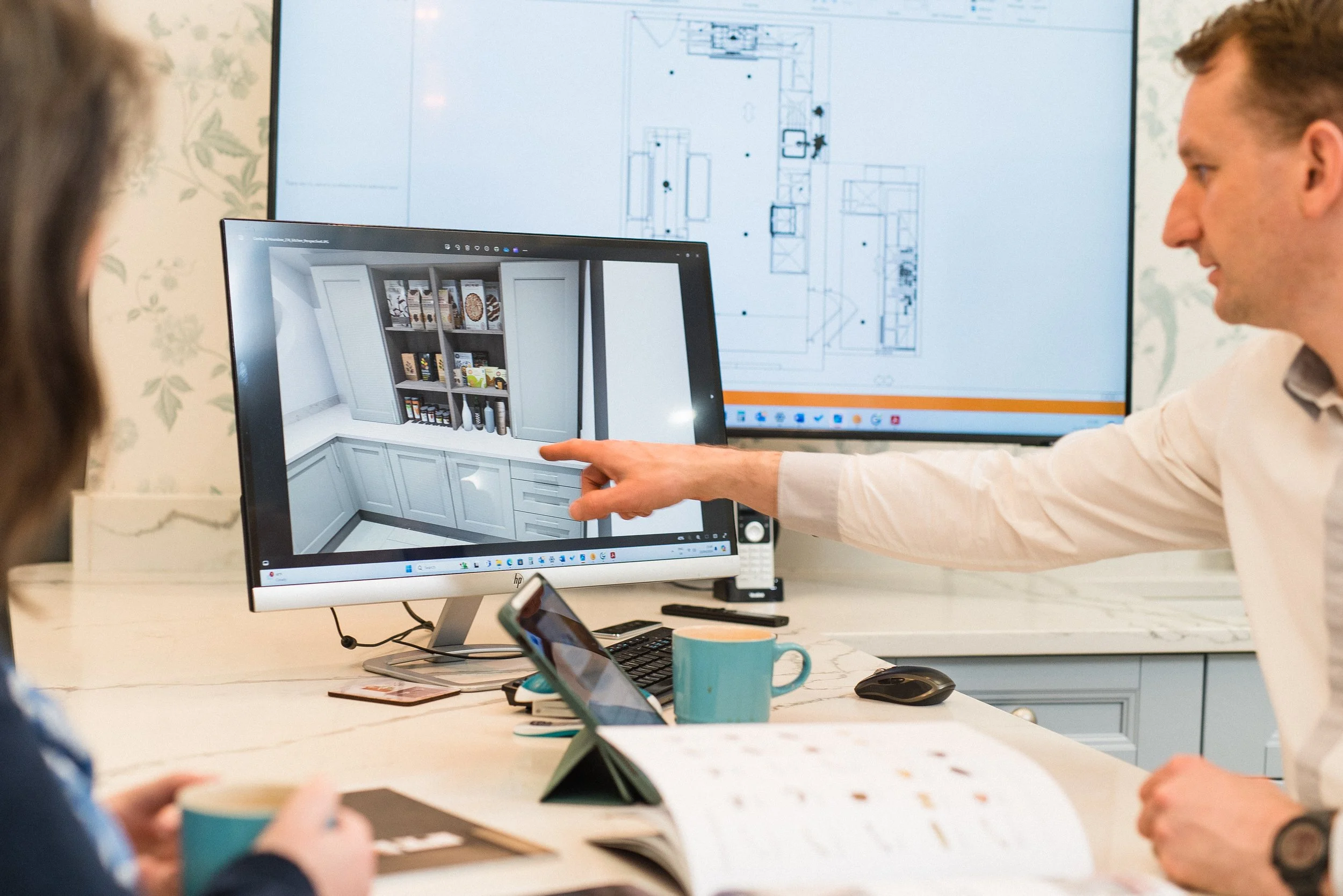 A man in a white shirt pointing at a 3D kitchen cabinet design on a computer monitor, while two other people observe. There is a large blueprint on a screen behind him. The workspace has a white table with a coffee mug, smartphones, and notepads.