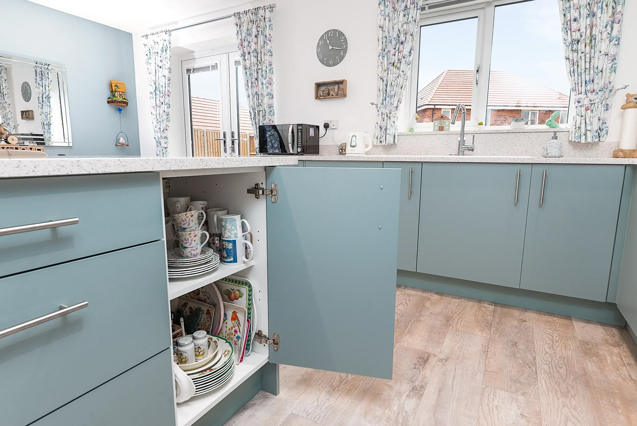 Kitchen with light blue cabinets, open cabinet showing dishes and mugs, window with floral curtains, stone countertop, clock on white wall, outside visible through window with houses and blue sky.