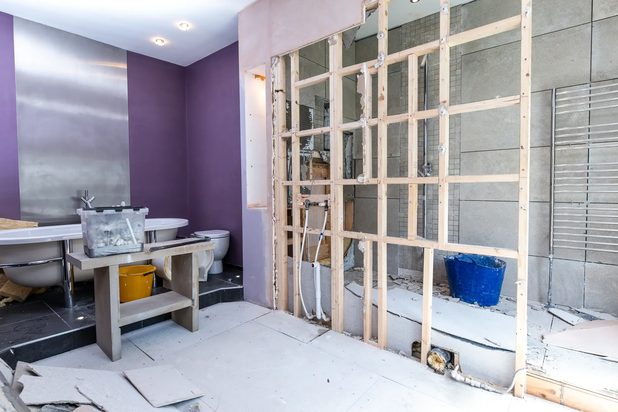 Bathroom undergoing renovation with a partially demolished wall, purple and metallic wall paint, a white bathtub, toilet, and construction tools and materials scattered around.