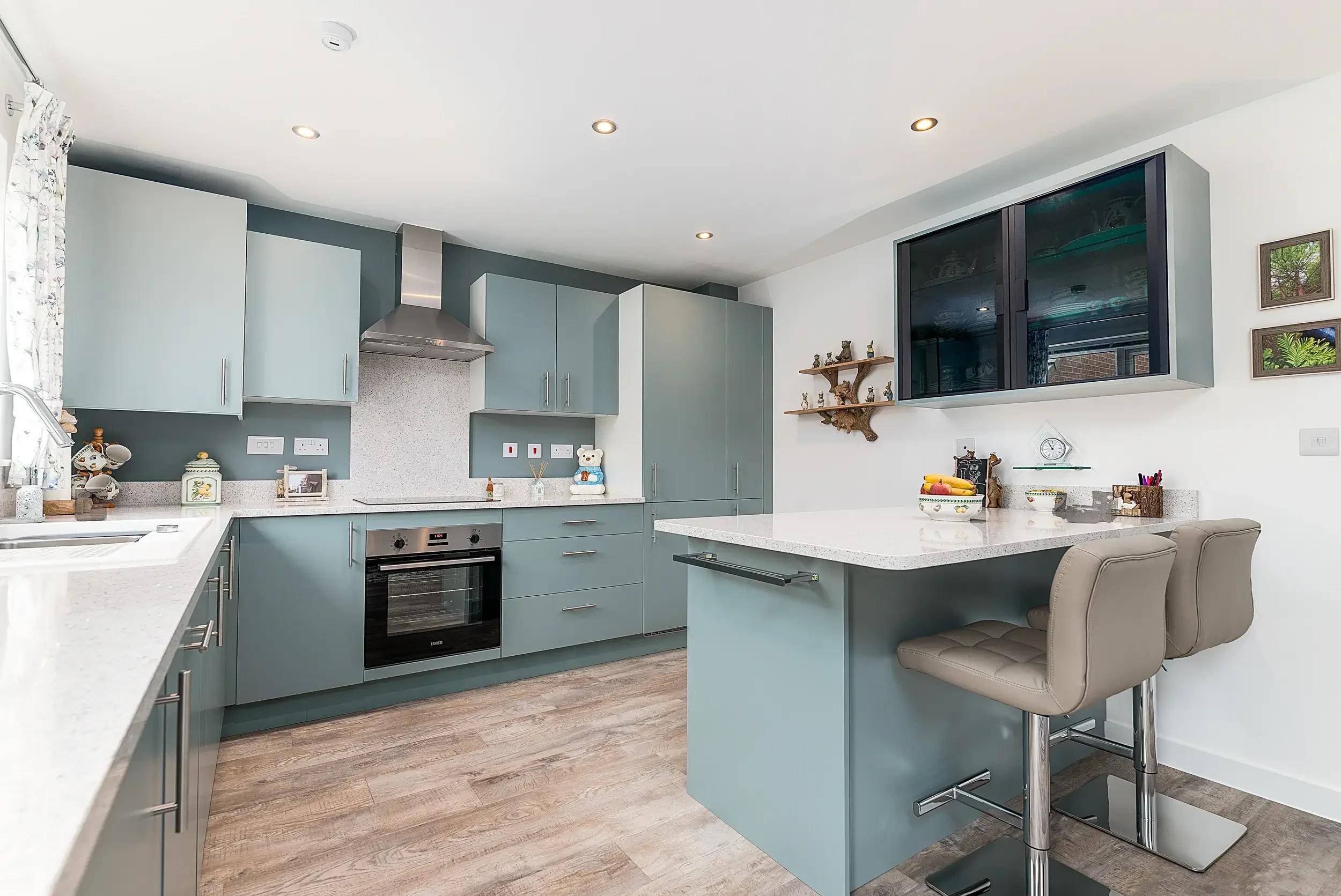 Modern kitchen with blue cabinets, white countertops, stainless steel oven, black windowed cabinets, light wood flooring, decorative items, and bar stools.