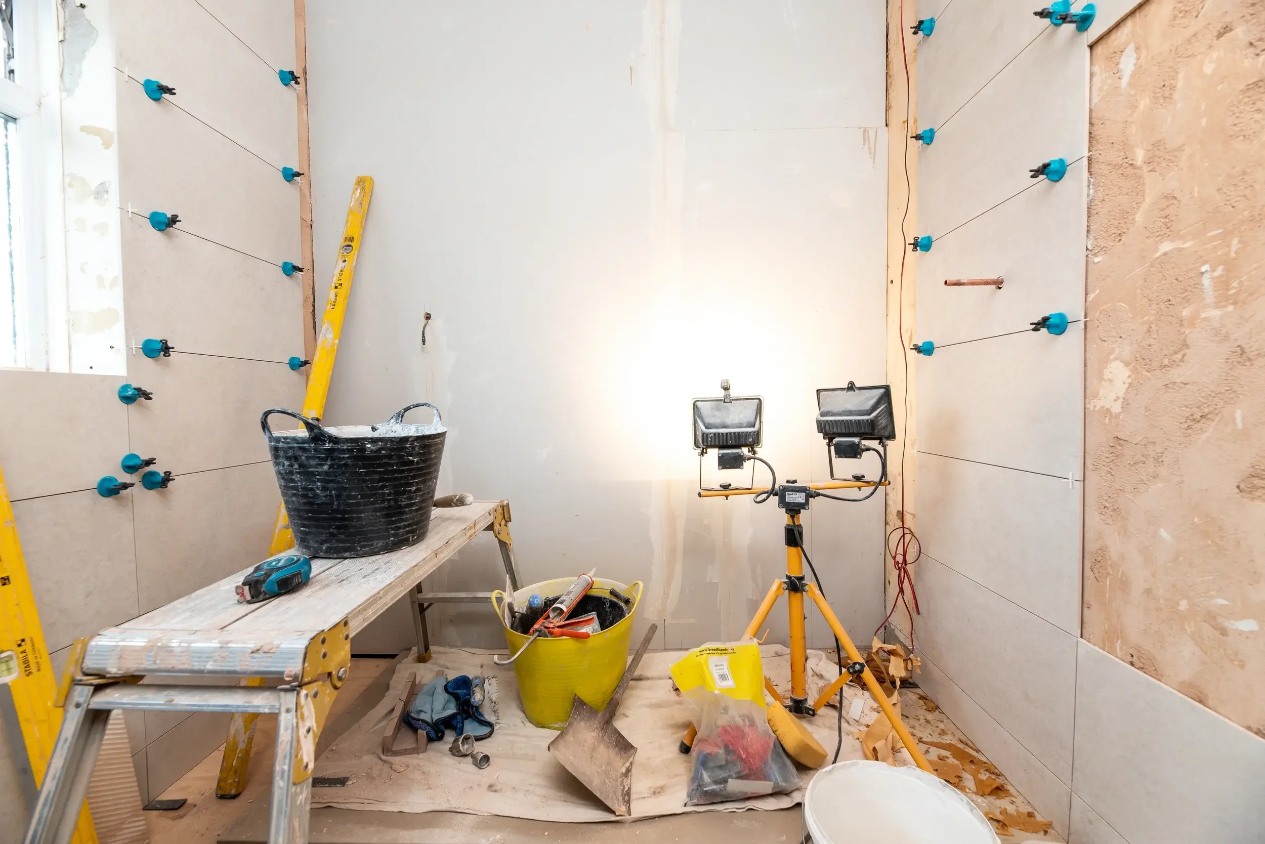 Construction site with tools and equipment, including a yellow ladder, a black bucket, a yellow bucket, light fixtures, and wiring, in a room under renovation