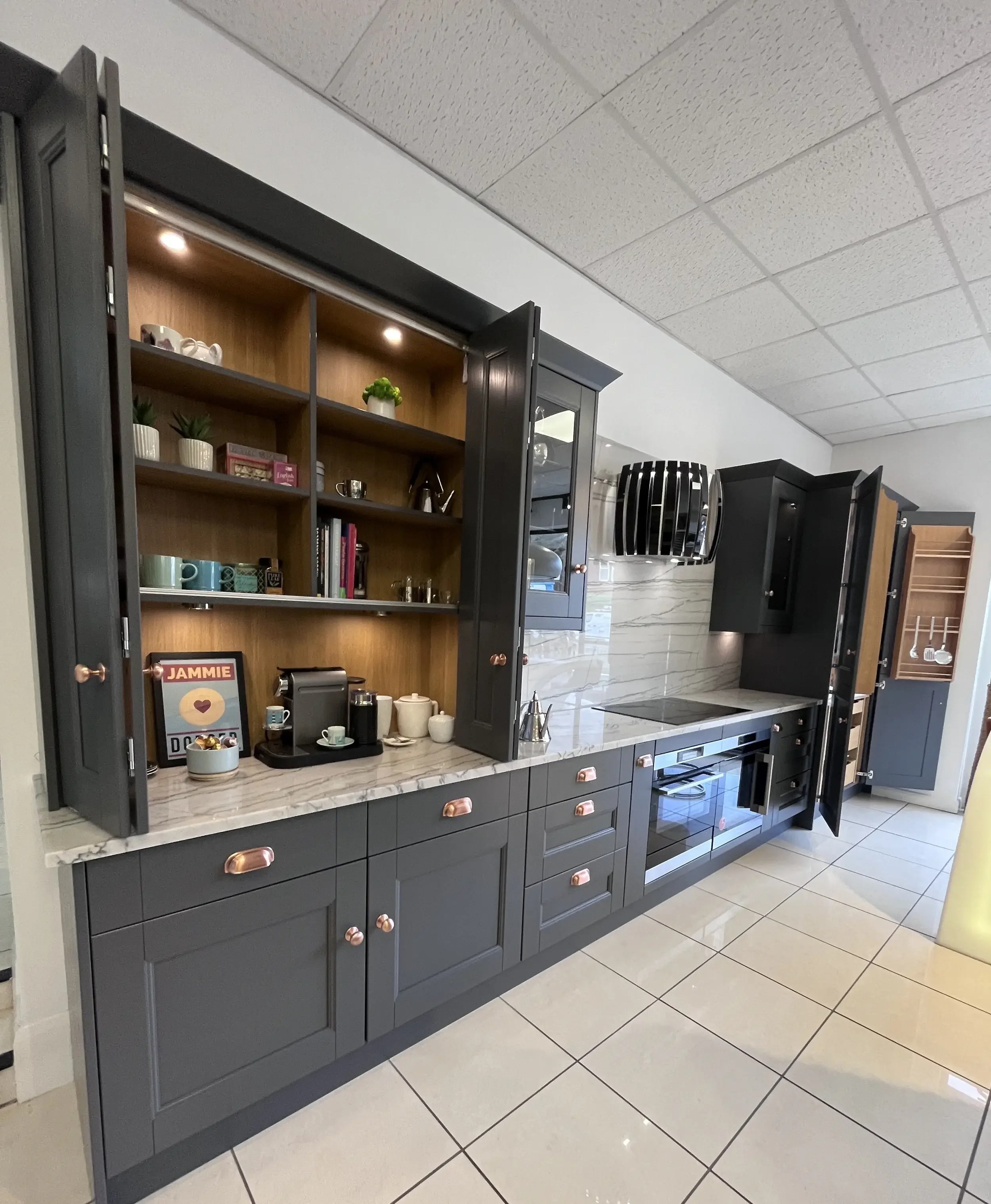 Kitchen with dark gray cabinetry, gold hardware, open shelves with cups, plants, books, and decor, built-in oven, white marble countertop, tiled backsplash, tiled floor, and ceiling tiles.