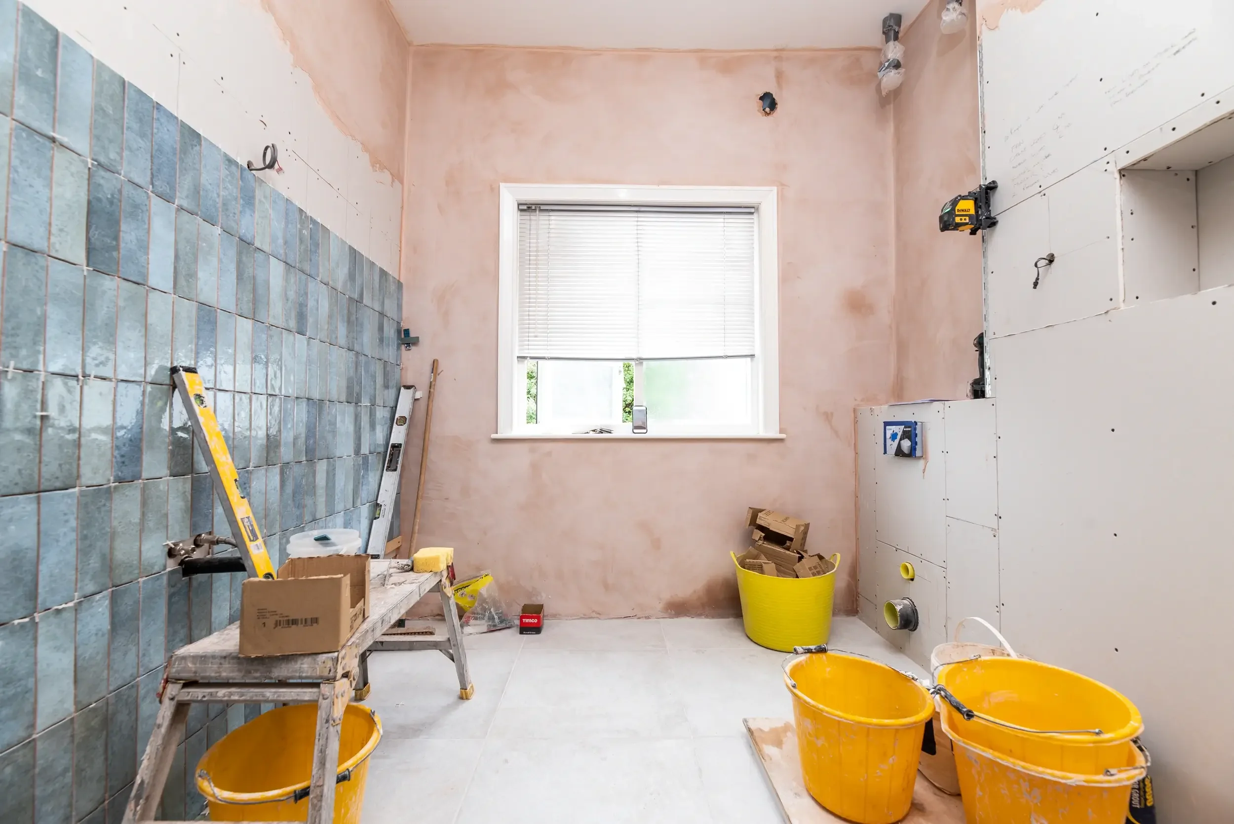 A room under renovation with partially tiled wall, construction tools, yellow buckets, and a window with blinds.