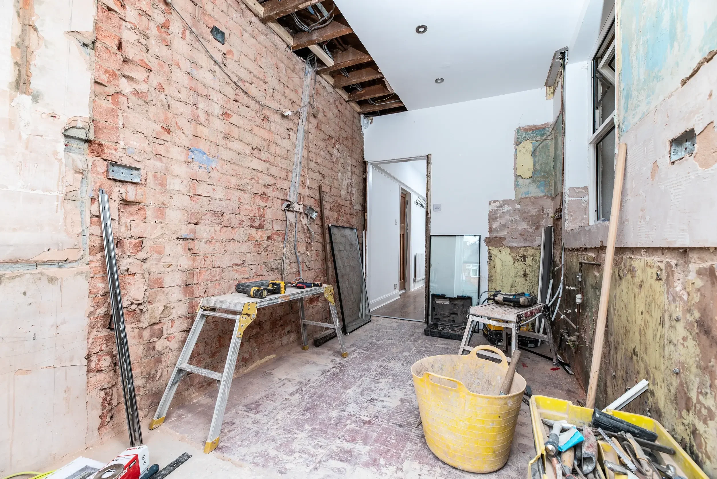 Room under renovation with exposed brick walls, construction tools, and supplies, including yellow buckets, ladders, and power tools. Part of the ceiling is open showing wooden beams and wires.