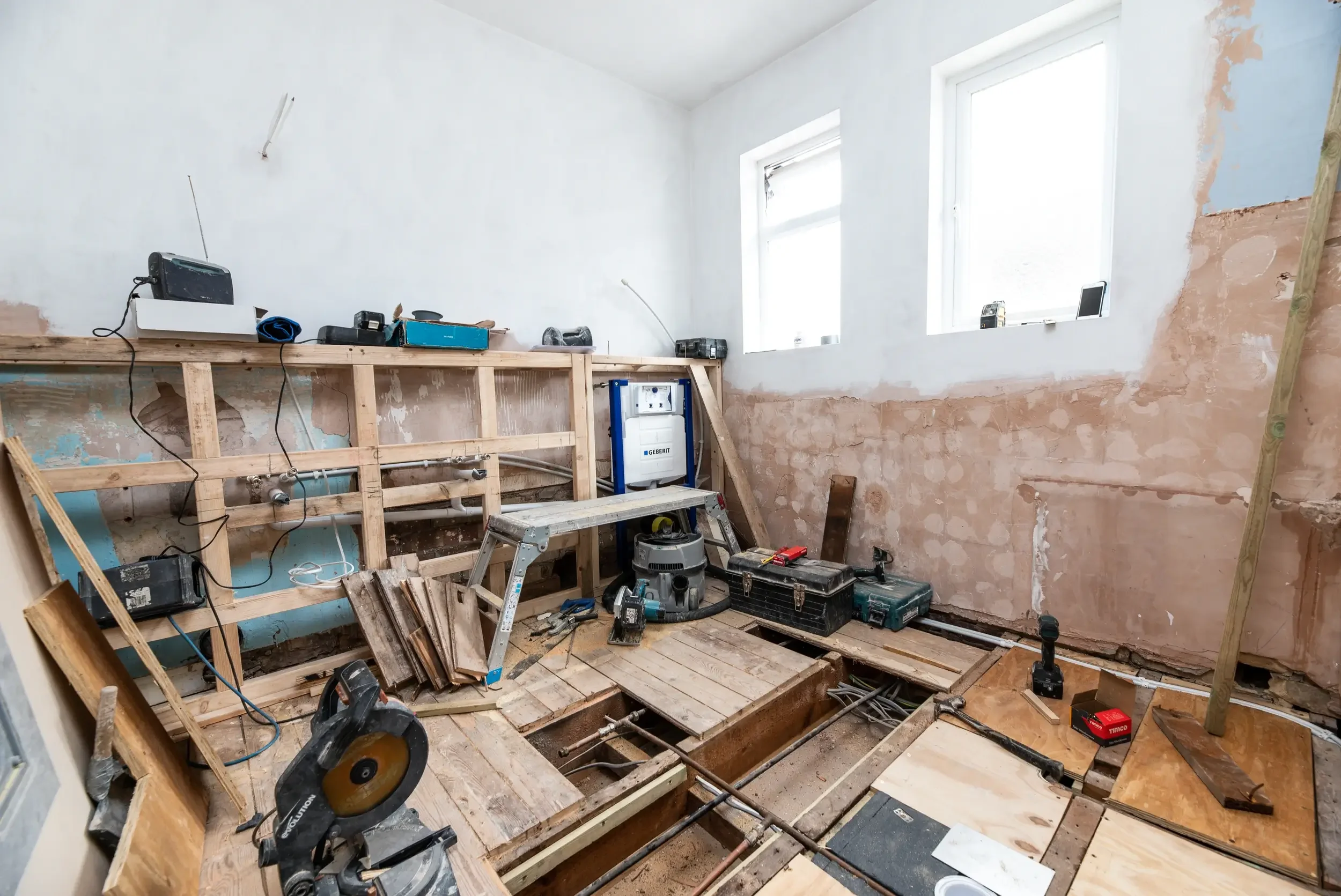 A room under renovation with partially built wooden framing, tools, power saws, drills, and construction materials scattered around. The walls are partially plastered, and there are two large windows letting in natural light.
