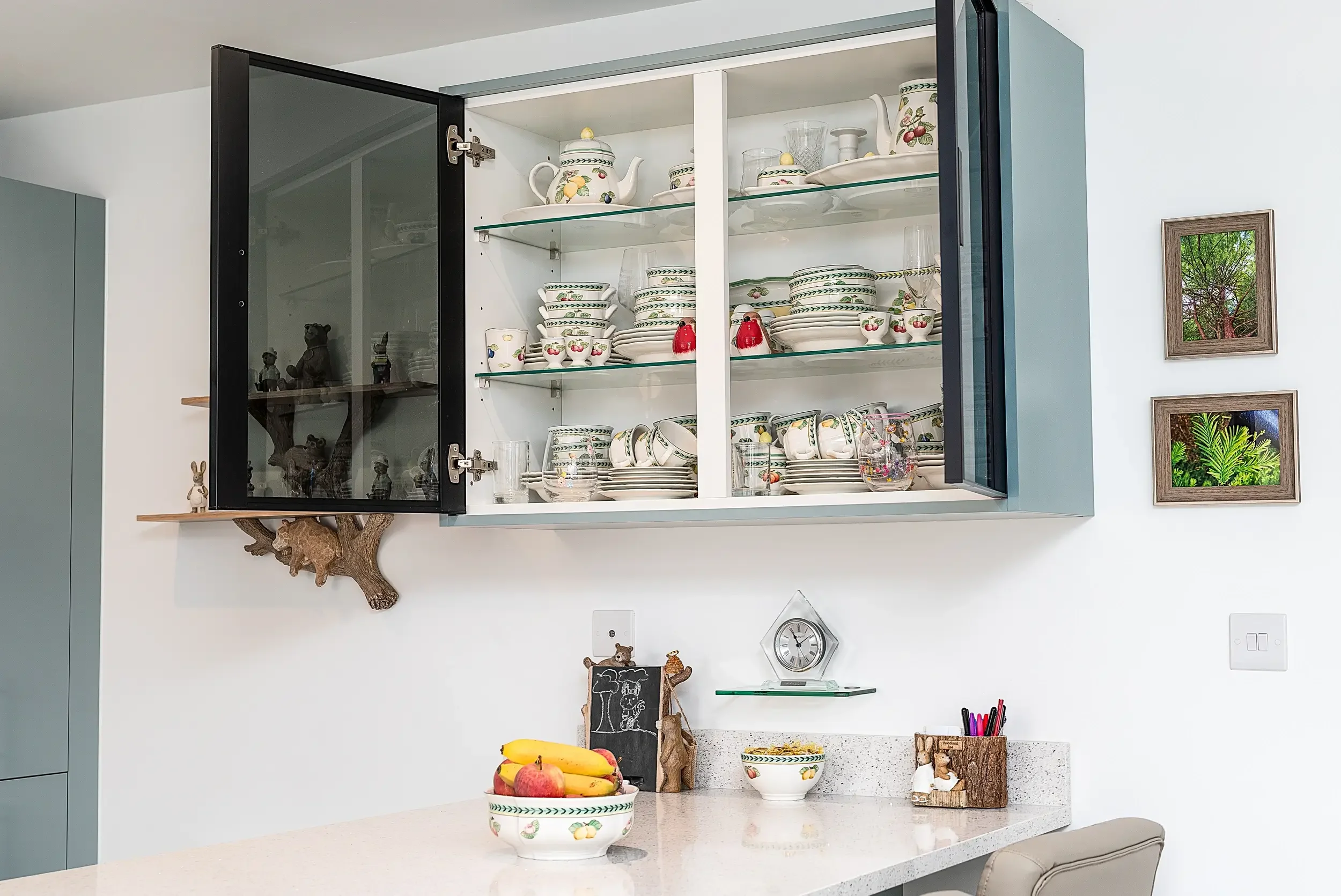 Kitchen with open upper cabinet filled with matching dishware including bowls, cups, and teapots with fruit-themed designs. On the countertop, there is a bowl of bananas and apples, a small chalkboard, and a decorative wood piece. The background cont