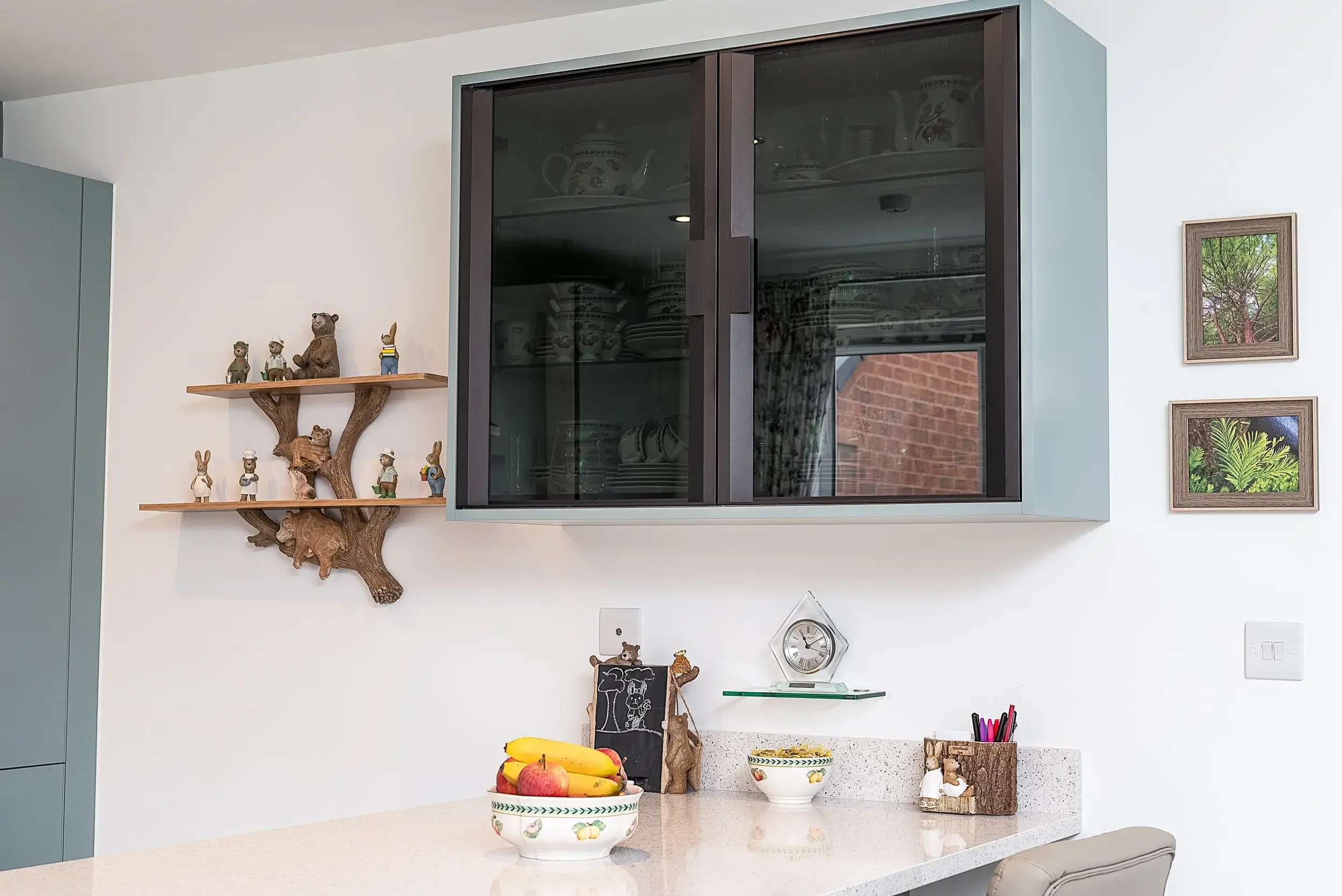 Kitchen with a white countertop, a bowl of bananas and apples, decorative figurines, a chalkboard, a clock, and framed pictures on the wall. Above the counter, a wall-mounted cabinet with glass doors displays dishes, and a wooden shelf with animal an