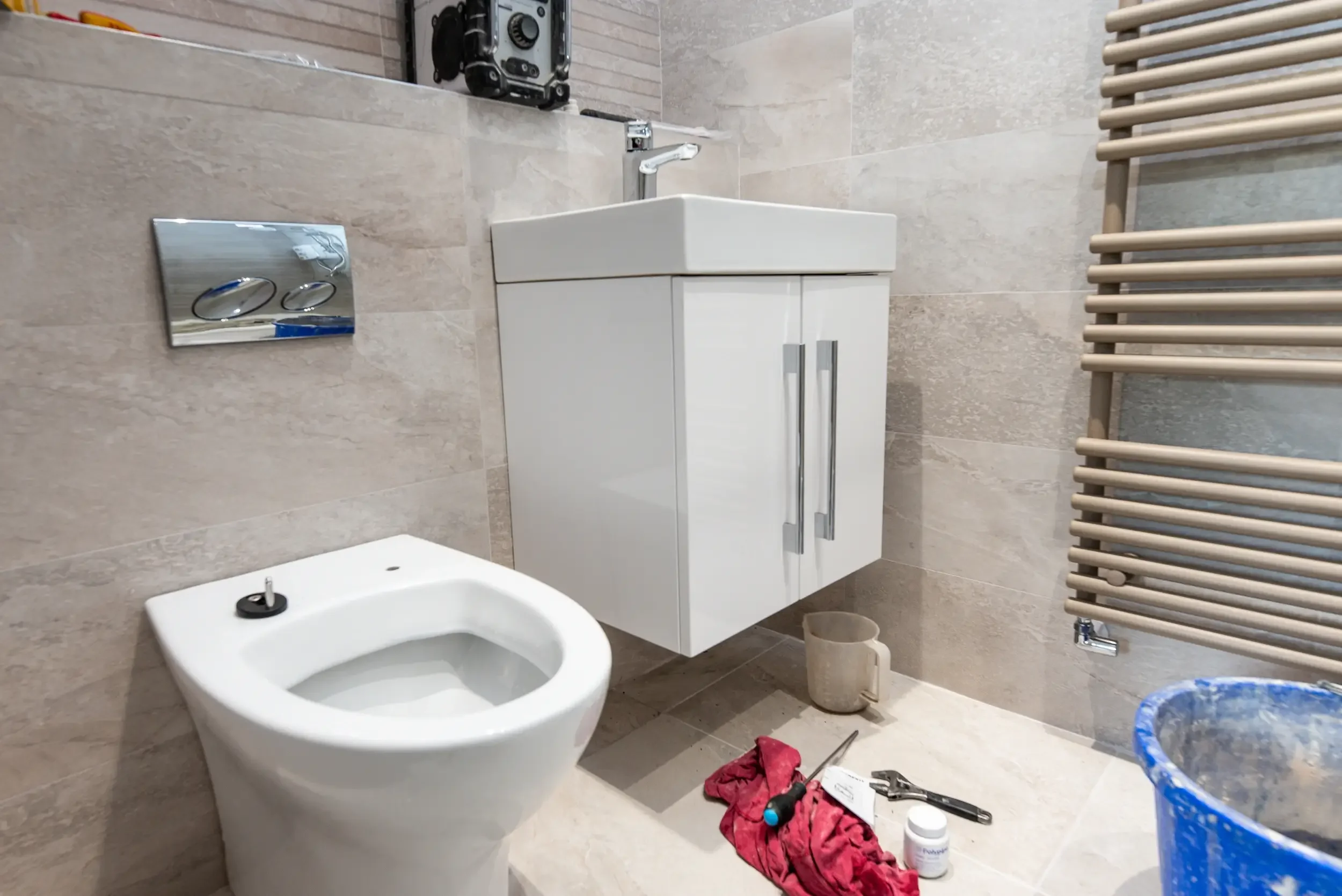 A partially renovated bathroom featuring a white toilet, a small white cabinet above it, and a bidet nearby. There are construction tools, red cloth, and cleaning supplies scattered on the floor. The walls are tiled with beige tiles, and a modern hea