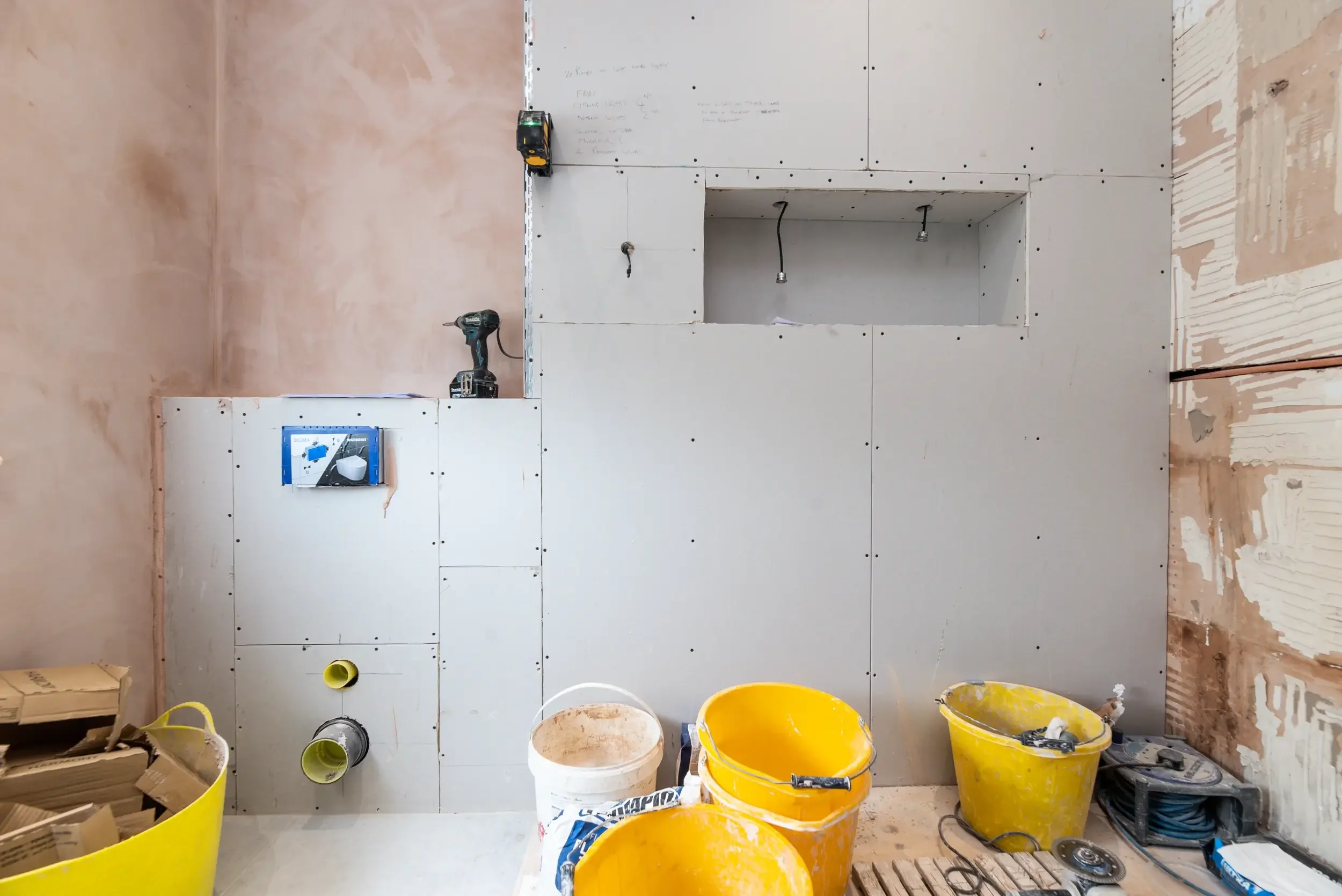 Construction site with partially finished drywall installation, with tools like drill and tape measure, and yellow buckets in the foreground.