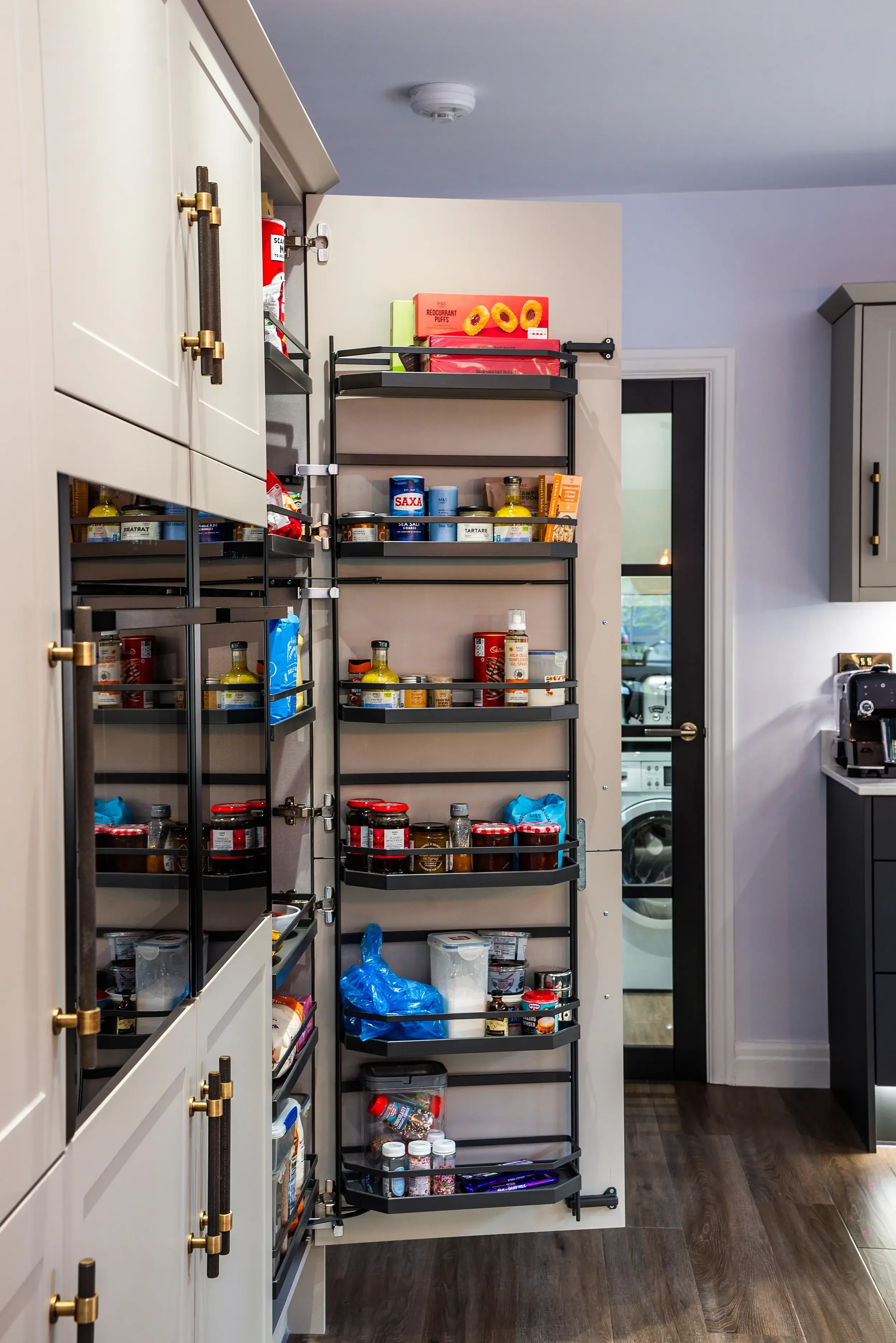 A pantry or kitchen storage area with a black rotating spice rack filled with jars, cans, and boxes, situated between white cabinets and near a door with a glass panel.