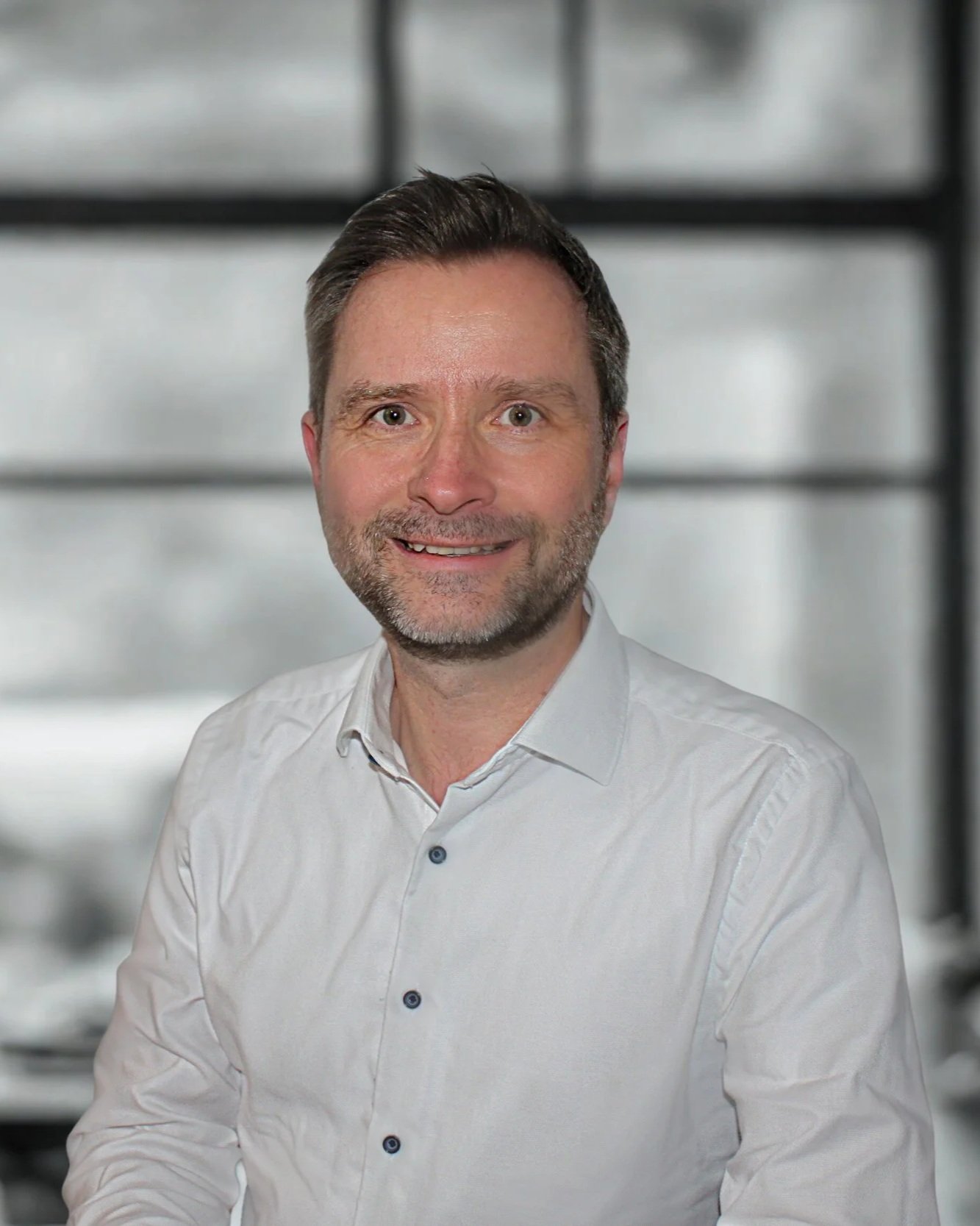 Porträt eines lachenden Mannes mit dunklem Haar und Bart, trägt ein weißes Hemd, im Hintergrund eine moderne, industrielle Raumgestaltung.