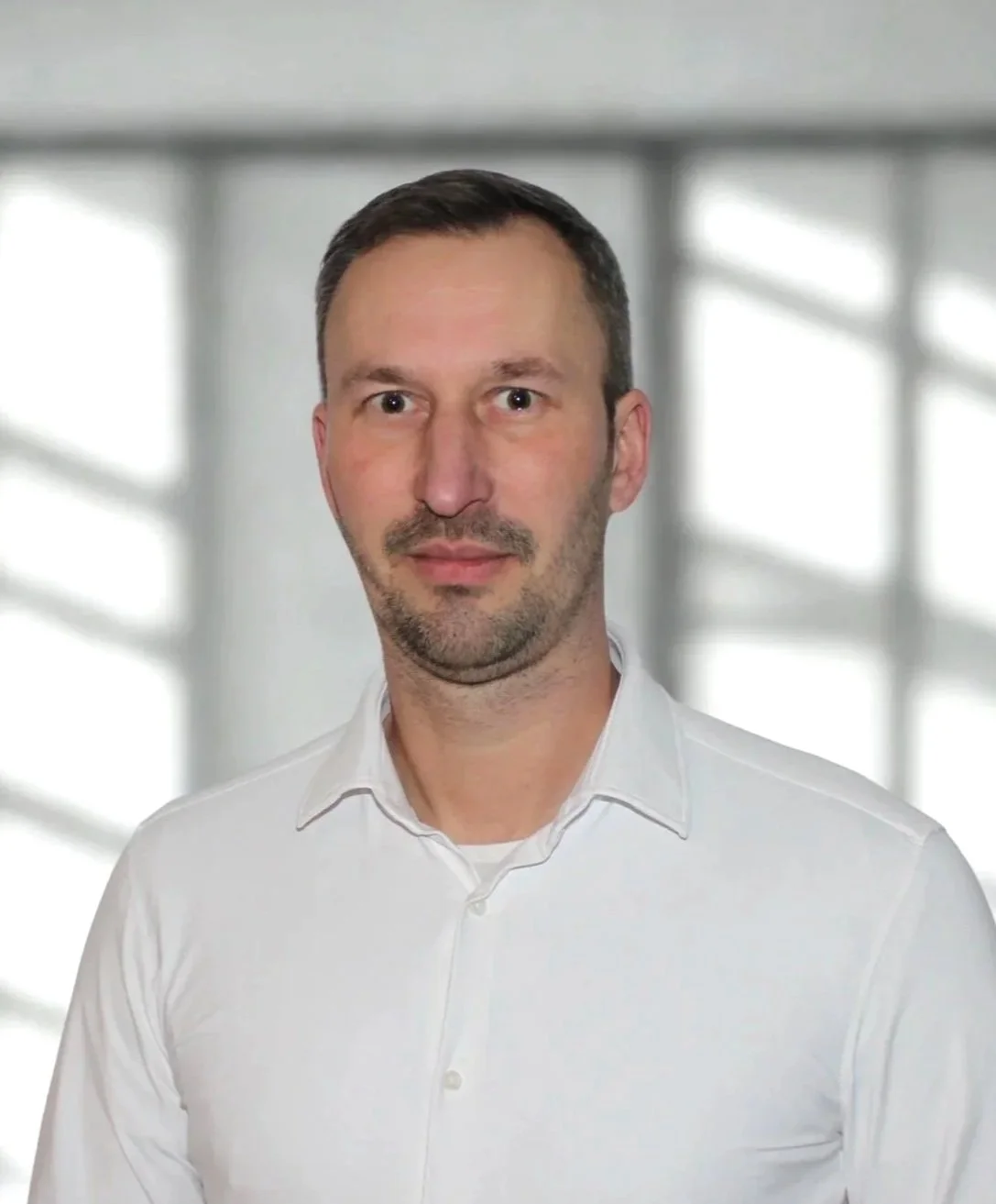 Porträt eines Mannes mit dunklem Haar, kurzen Bart, weißem Hemd vor unscharfem Hintergrund mit Fenstern.