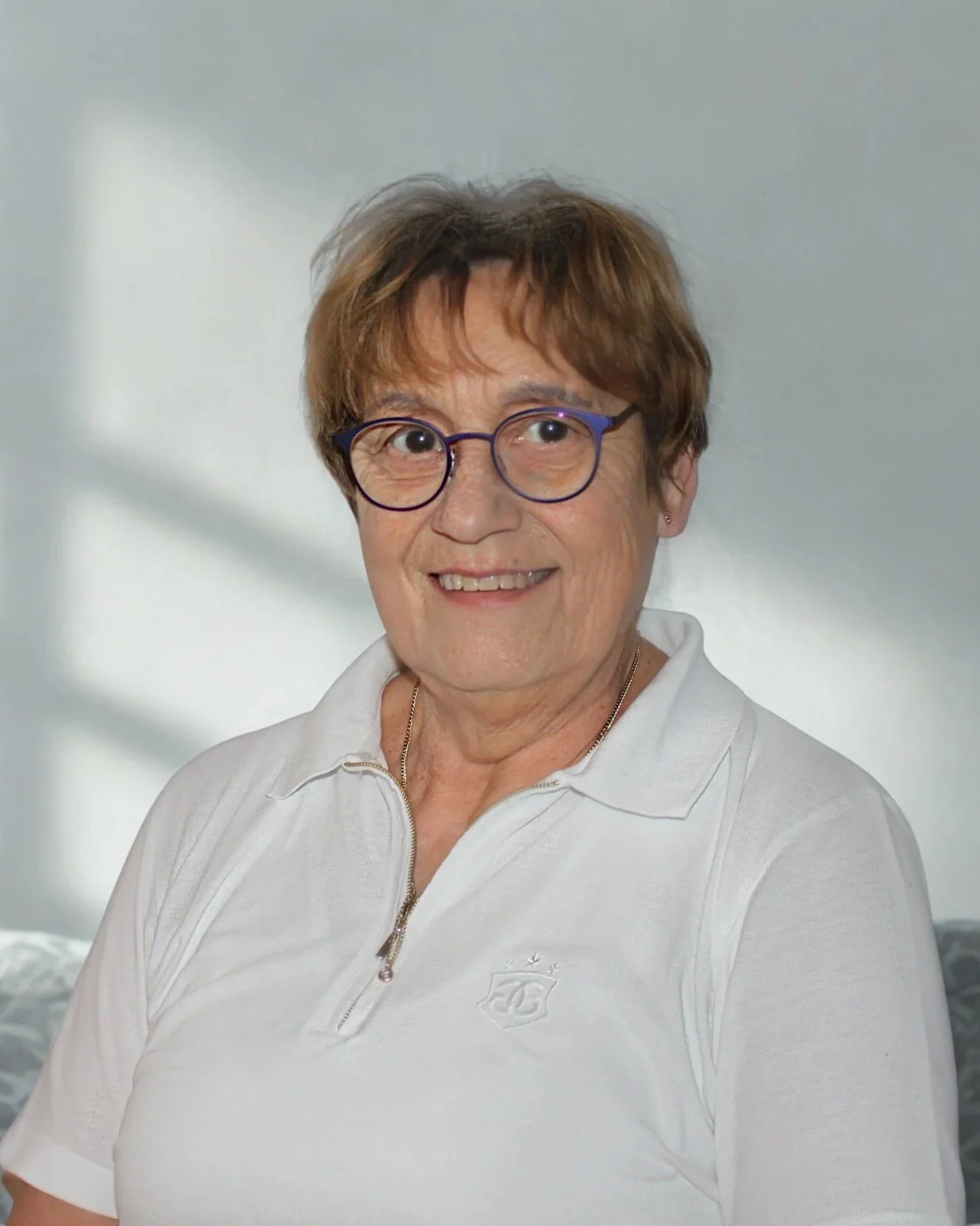 Eine ältere Frau mit kurzen braunen Haaren, Brille mit lila Rahmen, trägt ein weißes Polo-Shirt, lächelt freundlich, im Hintergrund eine helle Wand mit Schatten.