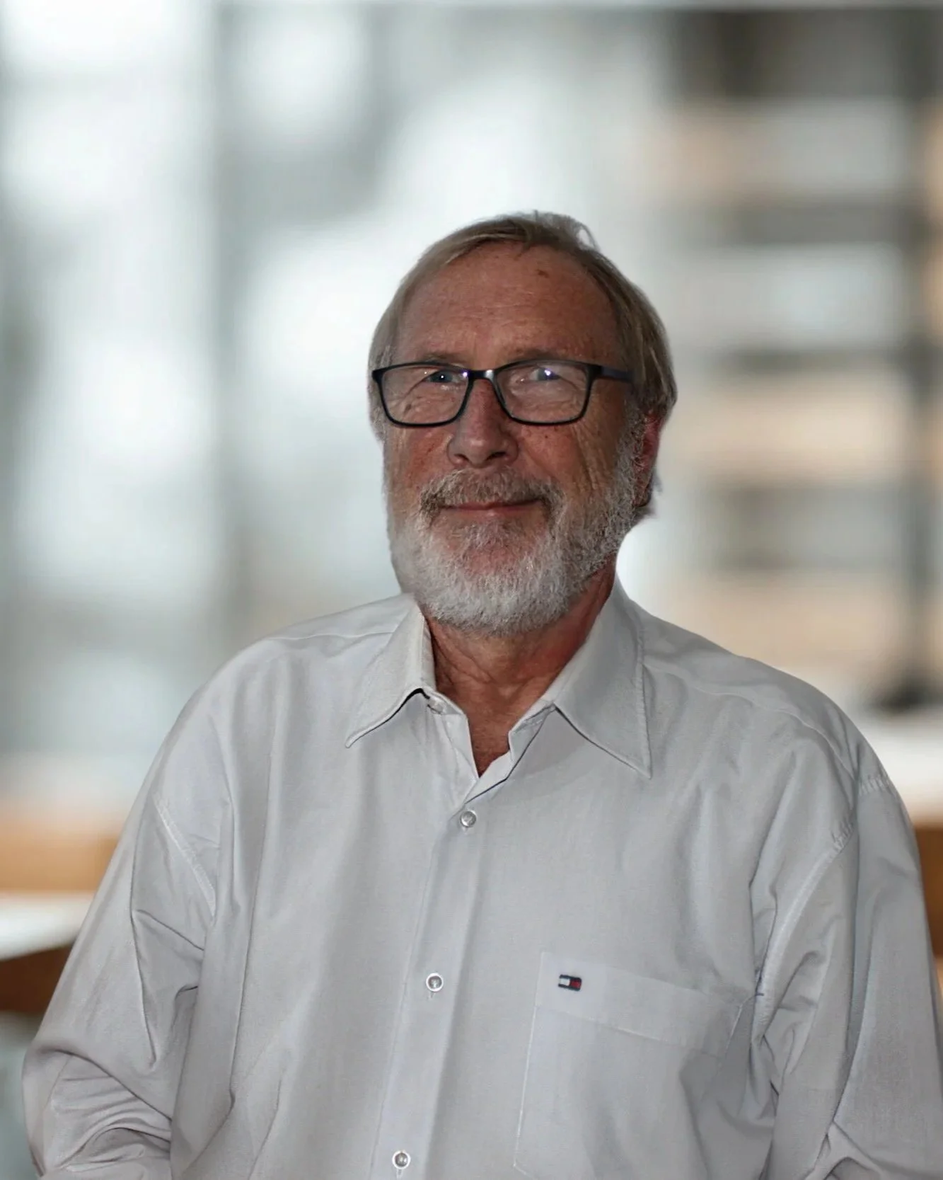 Älterer Mann mit Brille, grauem Bart und weißem Hemd in einem Raum mit unscharfem Hintergrund.