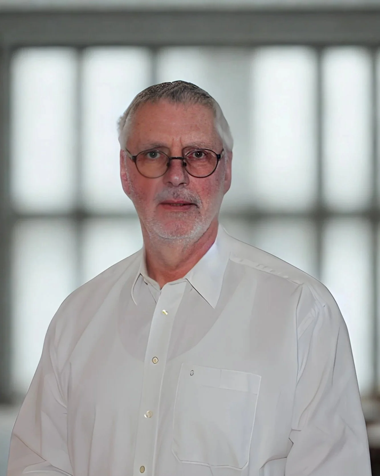 Ein älterer Mann mit Brille und weißen Haaren trägt ein weißes Hemd und steht vor einer großen Fensterfront mit quadratischen Mustern.