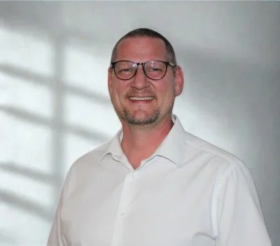 Ein Mann mit Brille in einem weißen Hemd, lächelnd, vor einem neutralen Hintergrund.