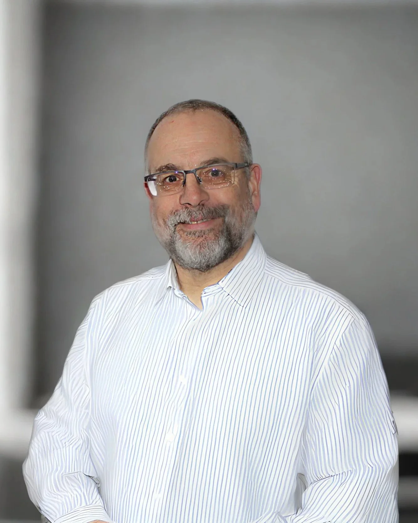 Porträt eines lächelnden Mannes mit Brille, grauem Bart und weißem gestreiftem Hemd vor grauem Hintergrund.
