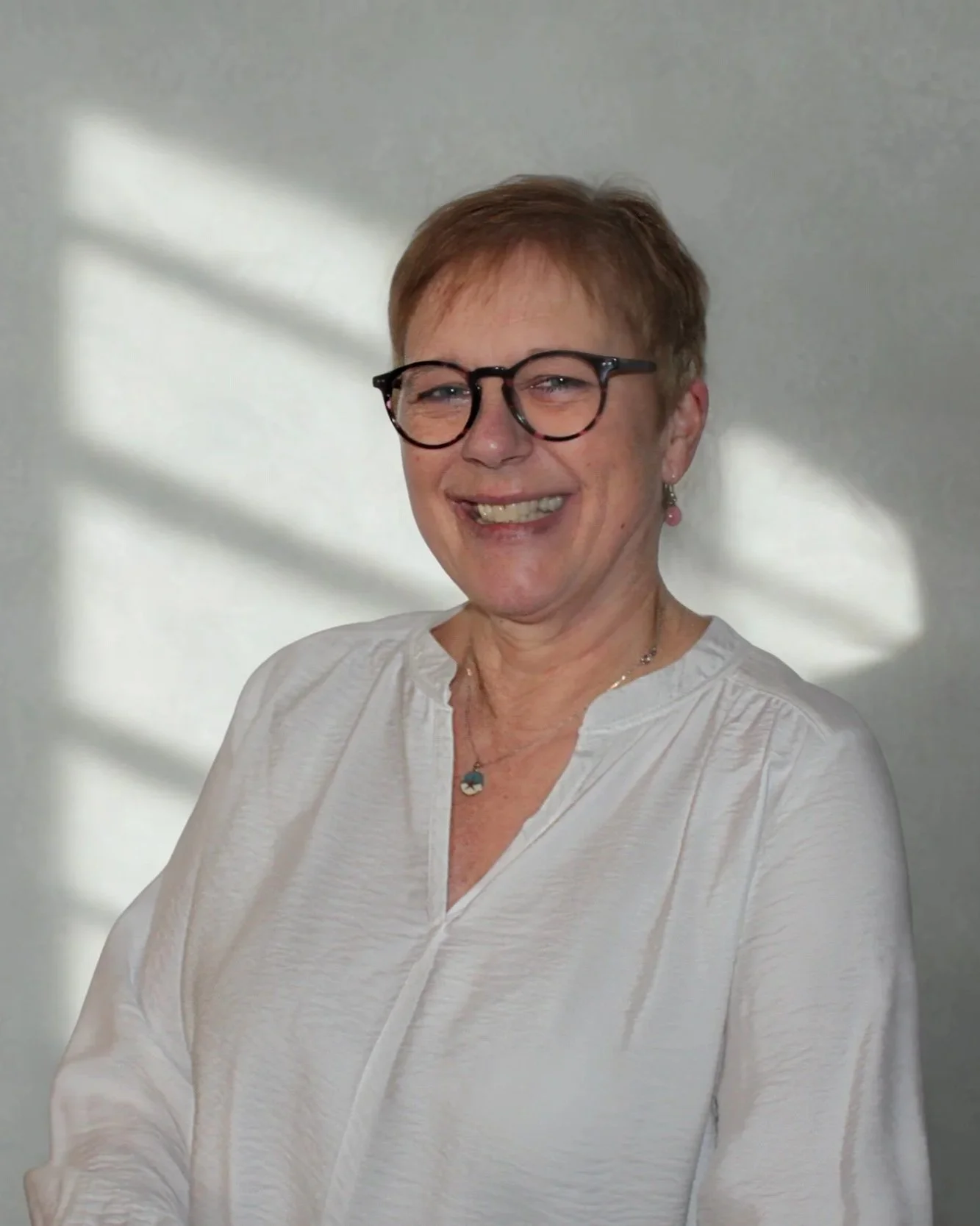 Lächelnde ältere Frau mit Brille, kurzem Haar und weißem Hemd vor einer grauen Wand.