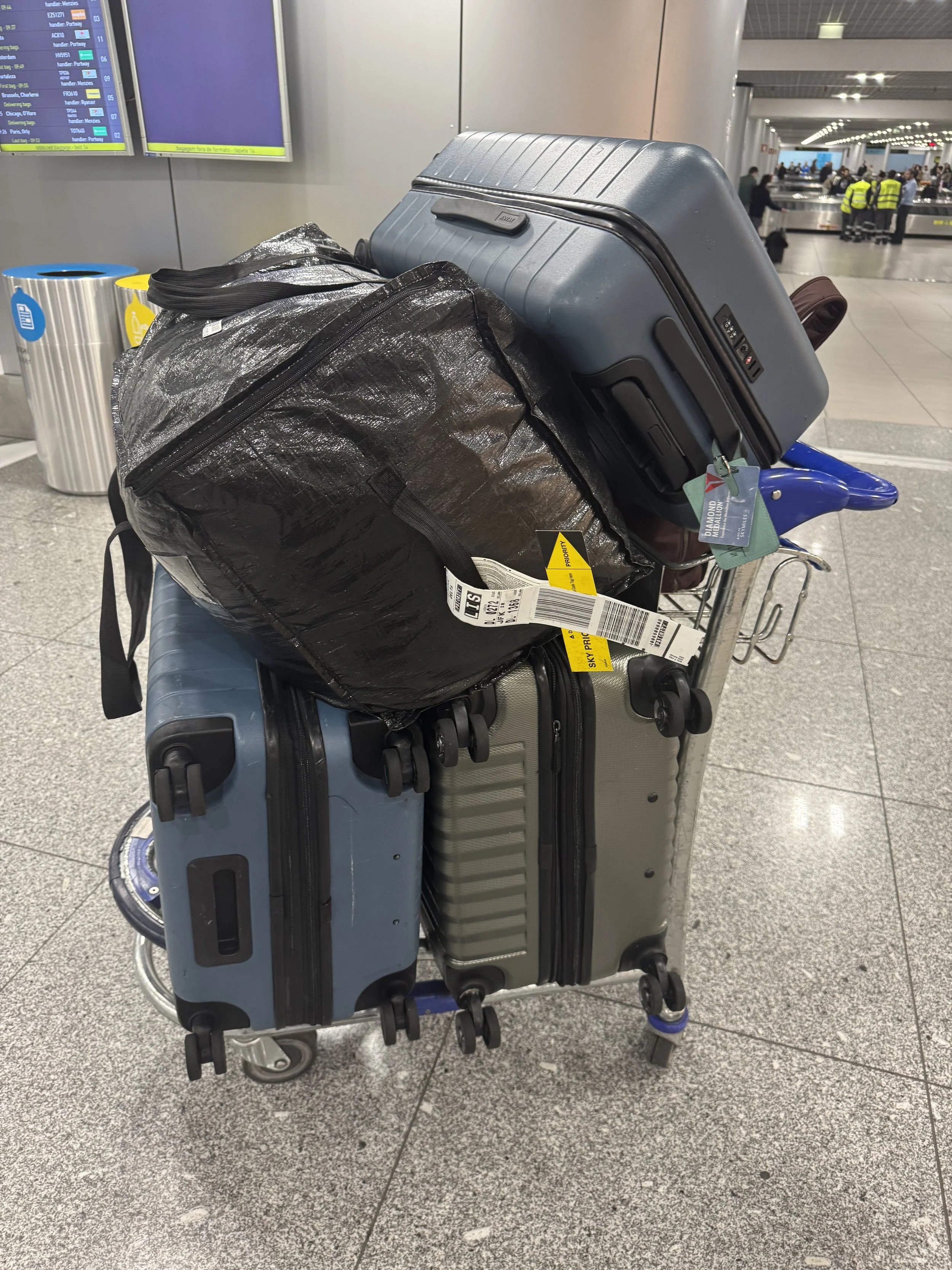A luggage cart at an airport stacked high with multiple large suitcases and bags. The bags are tightly packed, suggesting a long-term move rather than a short trip. A moment of returning with more belongings than before.