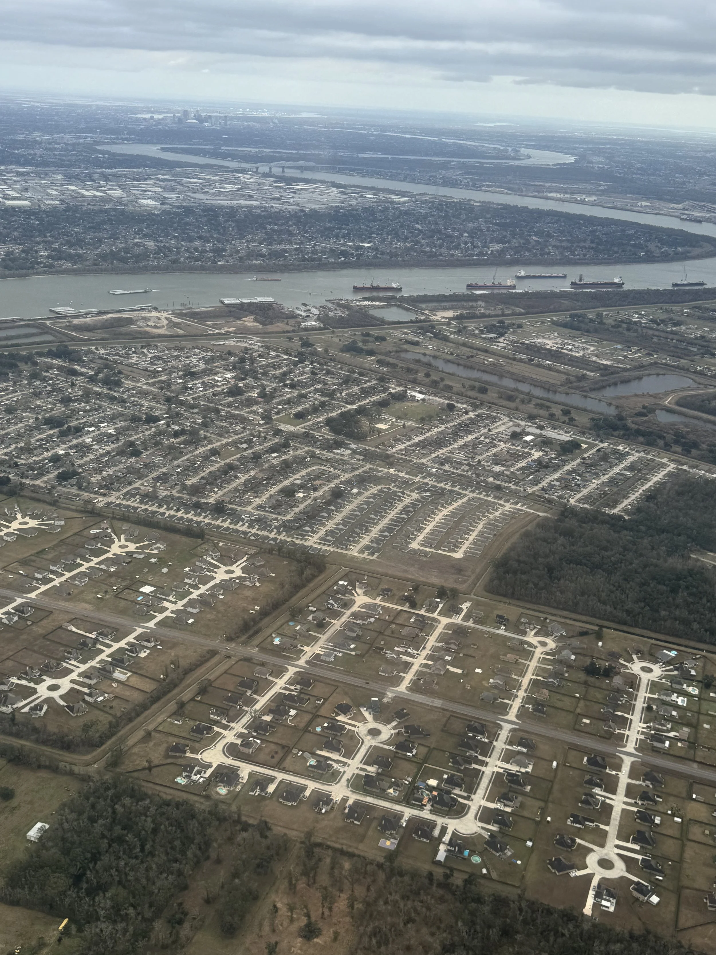 Aerial view of New Orleans from an airplane window, with the Mississippi River winding through the city below and ships visible along the water, seen from above during daylight.