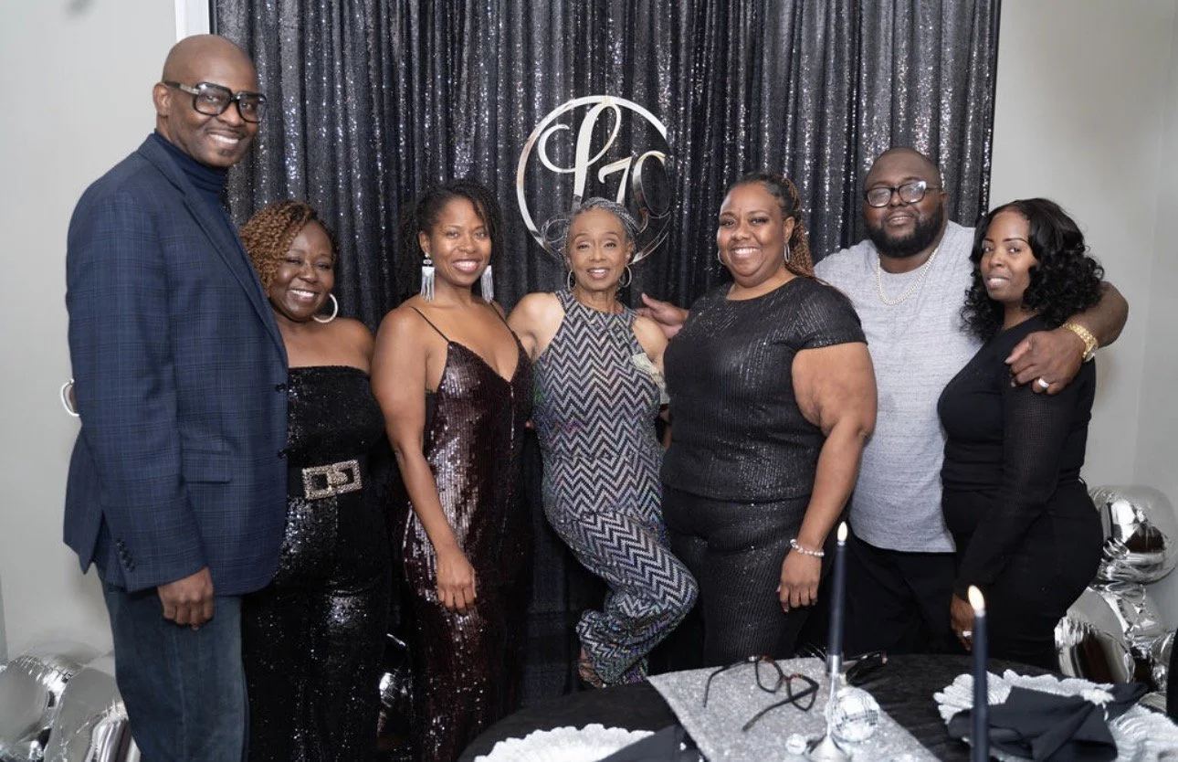 A group photo of Jacinta with her siblings and their partners at her mom's 70th birthday celebration.  Everyone is dressed in black and metallic tones, standing closely together and smiling in front of a decorated backdrop.