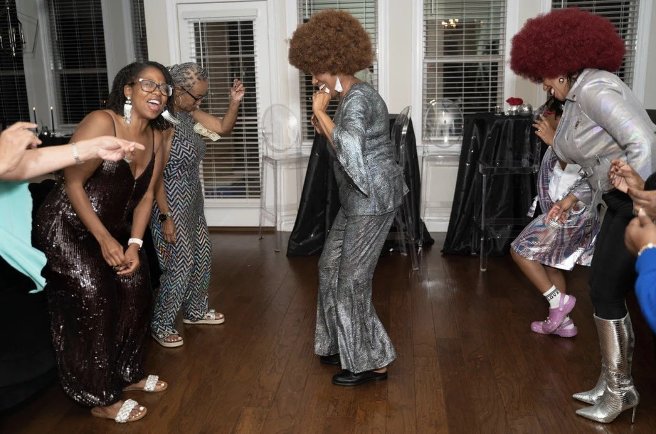 A multi-generational group of Black women dancing joyfully in a line at a family celebration, with elders and younger relatives moving together.