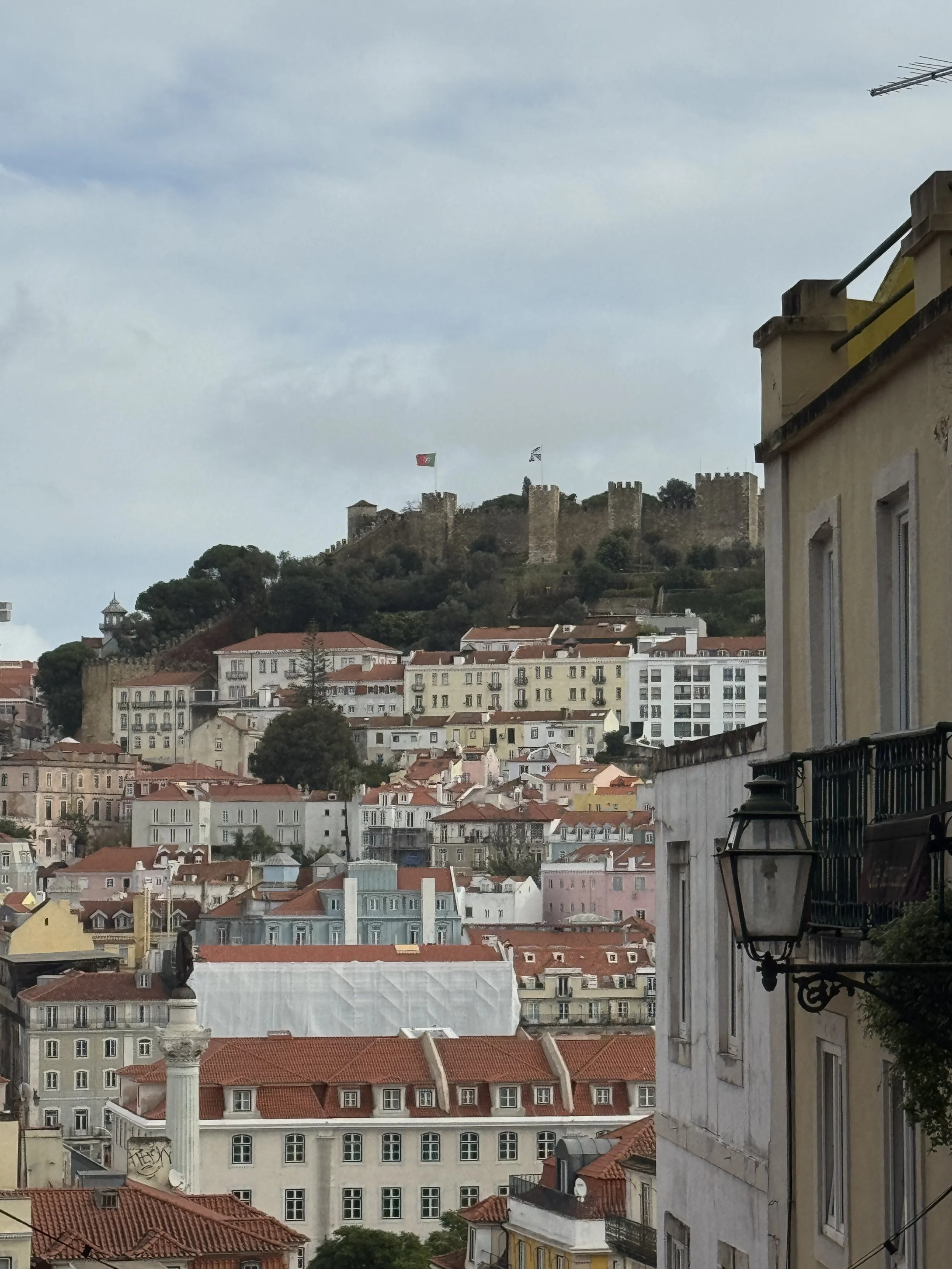 A view over Lisbon with red rooftops, pastel buildings, and São Jorge Castle in the distance under a cloudy sky. The image reflects looking back over a year of life in Lisbon and stepping into year two with more clarity.