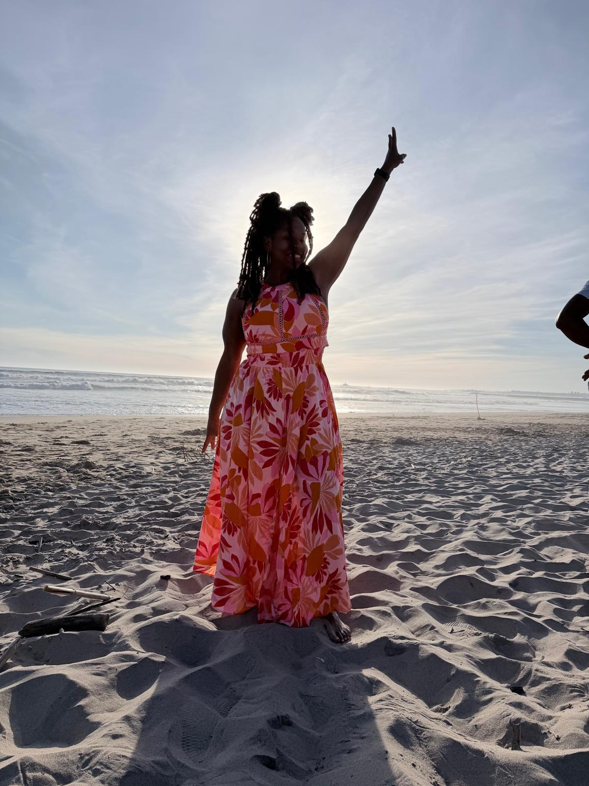 Jacinta stands barefoot on a beach near Lisbon, wearing a bright pink and orange dress with one arm raised toward the sky. The sun is behind her, creating a soft silhouette, with sand and the ocean in the background.