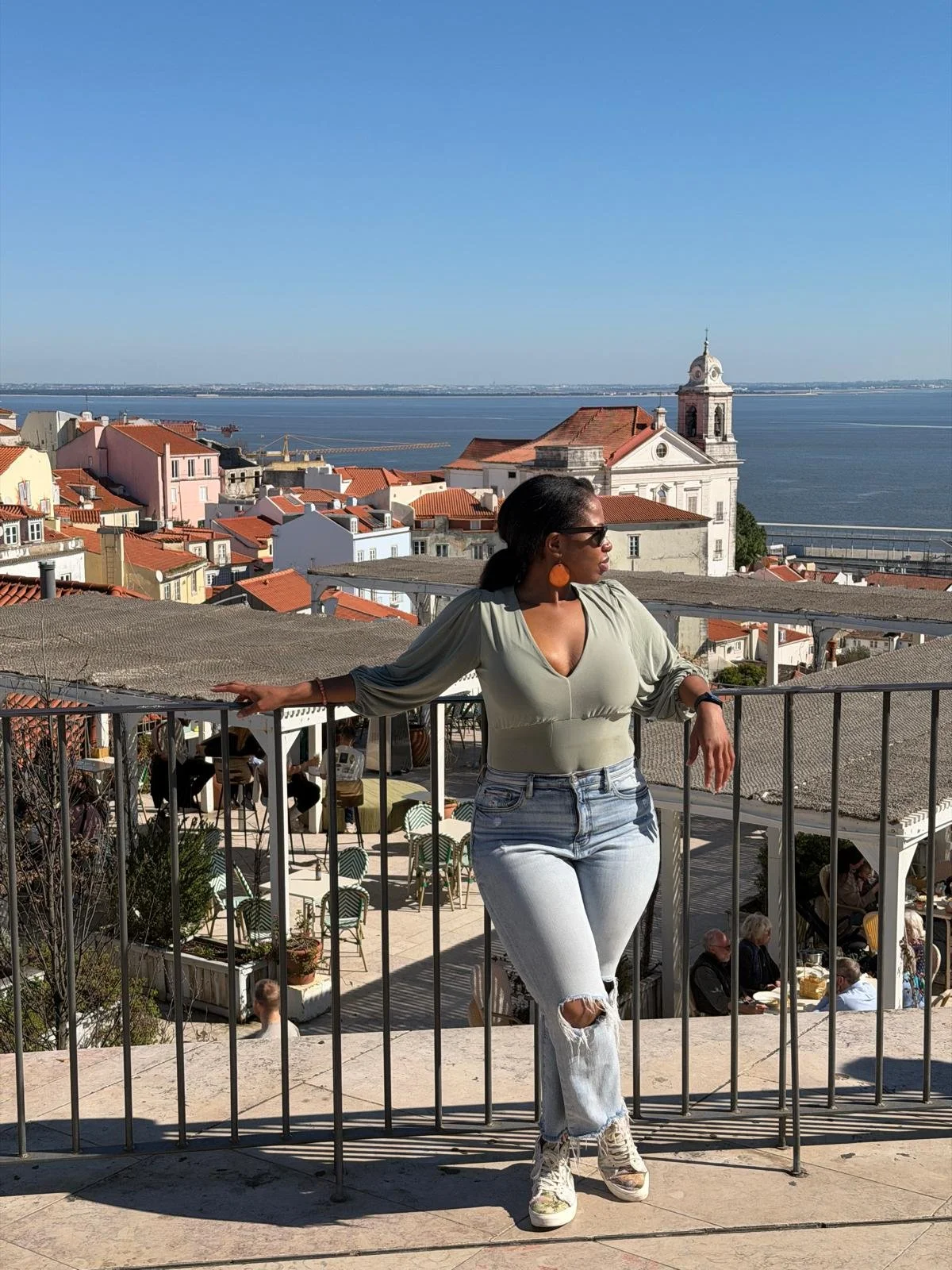 Jacinta stands on a scenic overlook in Lisbon, leaning against a railing with red rooftops, a church, and the Tagus River behind her. She is wearing sunglasses, jeans, and a green top, looking out across the city on a bright day.