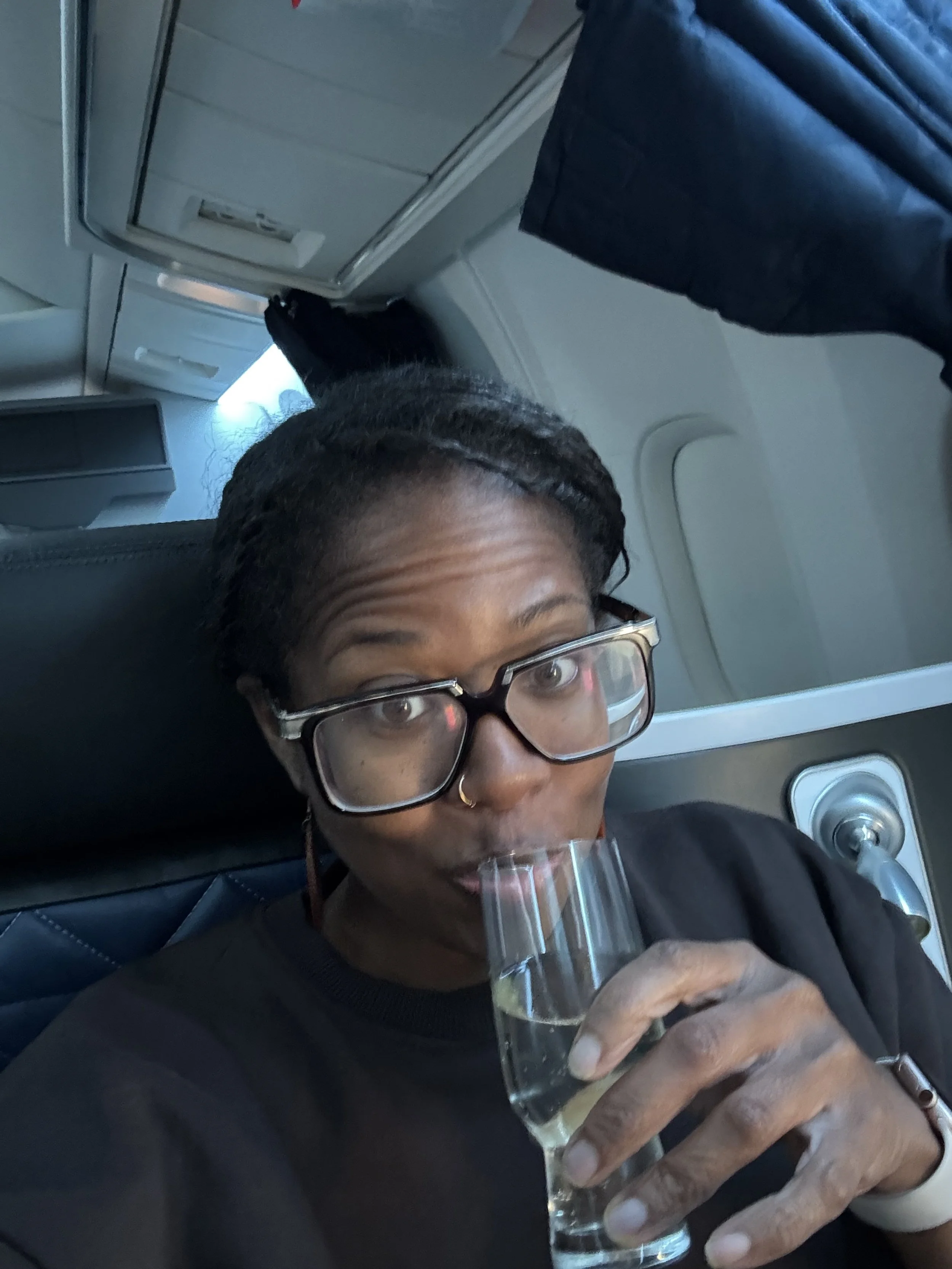 A woman wearing glasses sits in an airplane seat, smiling while sipping a glass of champagne, captured in a candid travel moment.