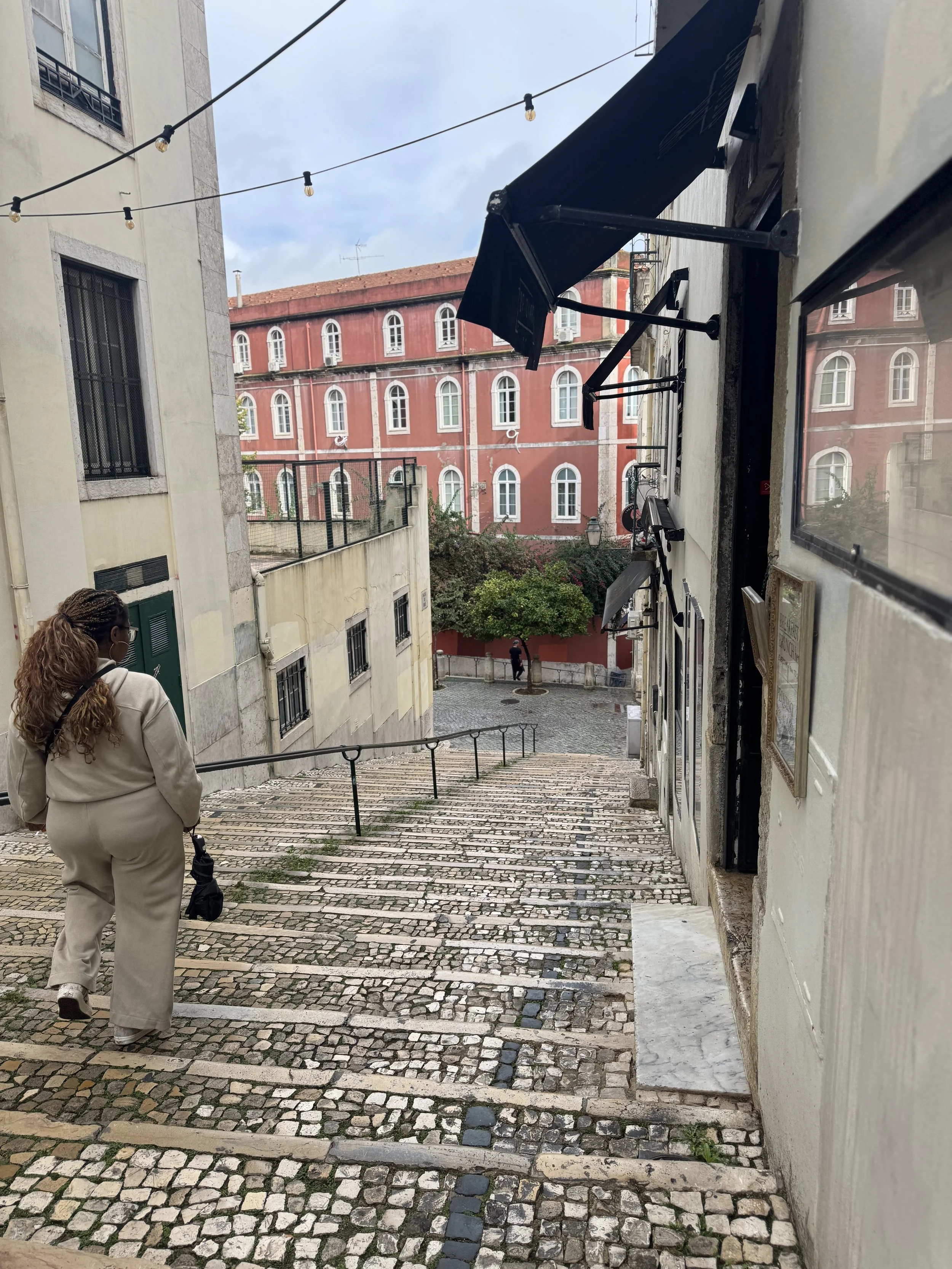 A street scene in Chiado, Lisbon, showing Jacinta walking down a sloped cobblestone street between tall buildings. The image reflects the neighborhood where Jacinta first landed in Lisbon and began finding her way into daily life there.