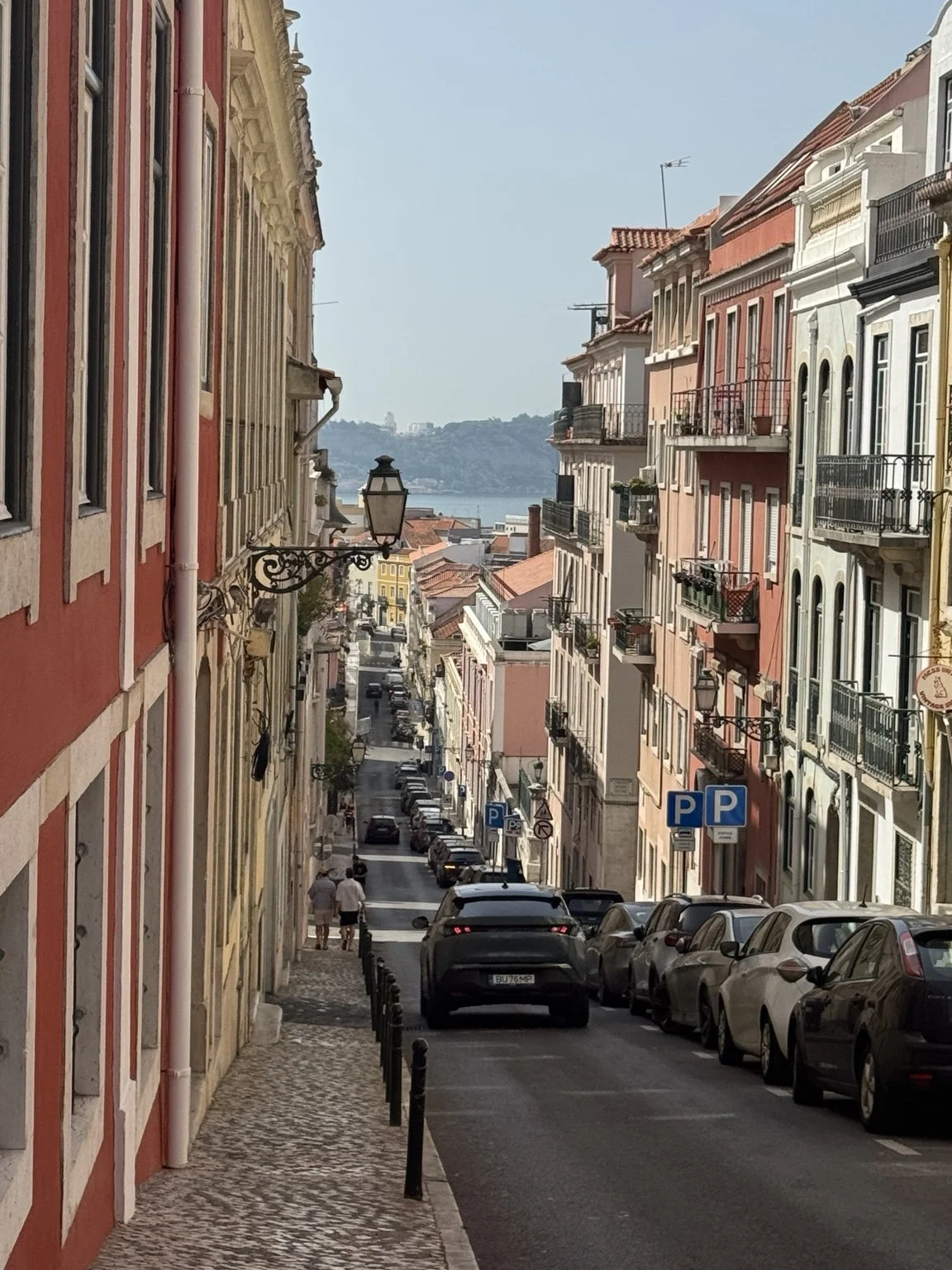 A sloped Lisbon street with colorful buildings, parked cars, and cobblestone sidewalks, with the Tagus River visible in the distance.