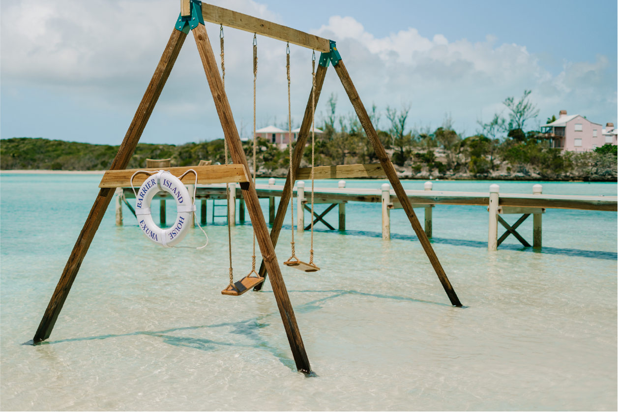 Empty swing set with a life buoy hanging on a sandy beach by turquoise water and a wooden pier, with houses and trees in the background.