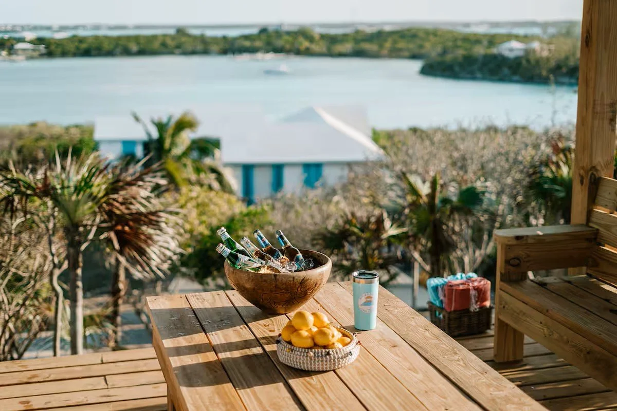 View of a wooden deck with a table holding a bowl of beer bottles, a basket of lemons, and a tumbler, overlooking a body of water with trees and buildings in the background.